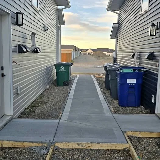 A narrow alleyway between two houses under construction, with a concrete sidewalk and three trash bins on the right side.