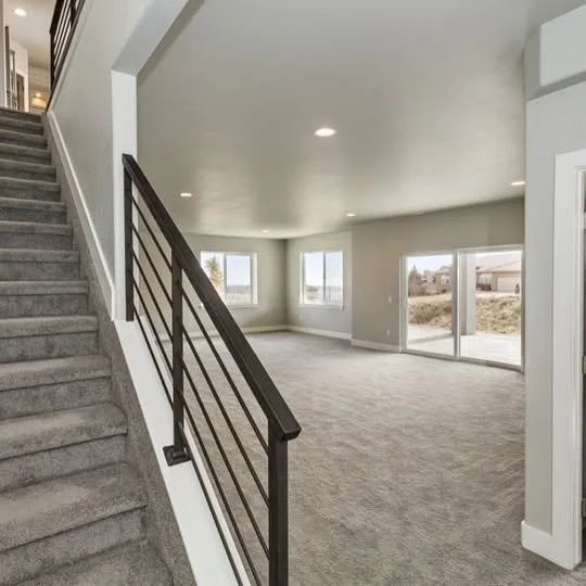 Empty living room with large windows, sliding glass door, carpeted floor, and staircase with metal and wood railing.