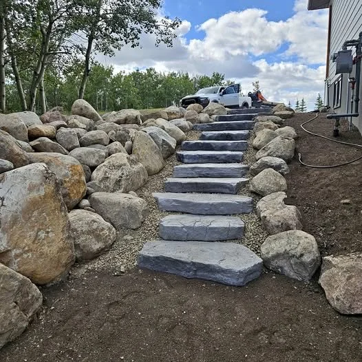 Stone steps leading up a slope with large rocks on each side, a white truck parked at the top, trees and a partly cloudy sky in the background.