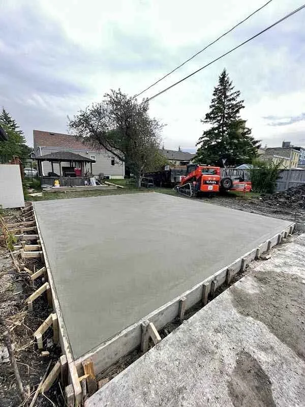 Freshly poured concrete slab foundation in a residential backyard with construction framing and equipment, including a mini excavator, under an overcast sky.