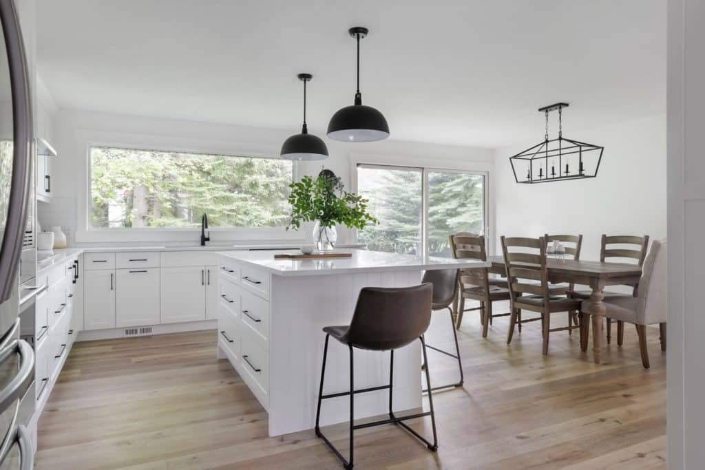 Bright modern kitchen with white cabinets, kitchen island, black pendant lights, wooden dining table with nine chairs, large window with view of trees, light wood flooring, and a plant on the island.