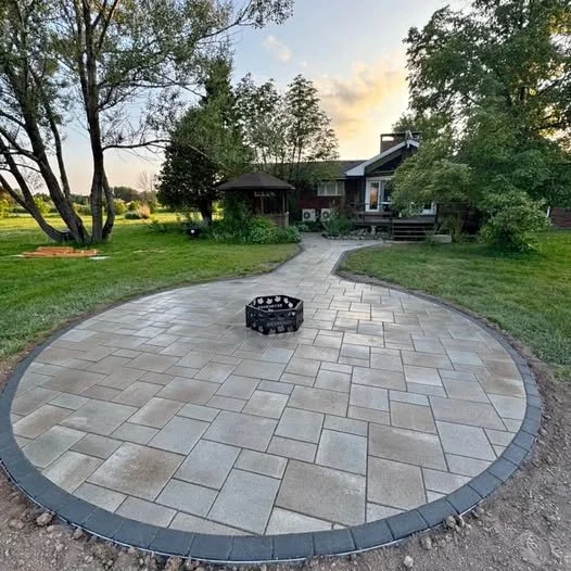 A curved stone patio with a fire pit in the center, surrounded by a lush yard with trees and a house in the background, during sunset.