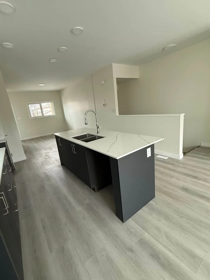 Modern kitchen with an island featuring a white marble countertop and black cabinetry, stainless steel sink, and light wood flooring in an open, bright space.