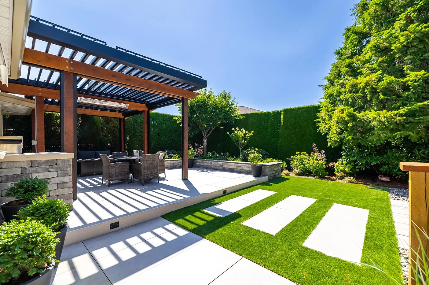 Modern backyard patio with a shaded pergola, outdoor dining table and chairs, potted plants, manicured grass, garden bed with shrubs and trees, and a tall hedge wall.
