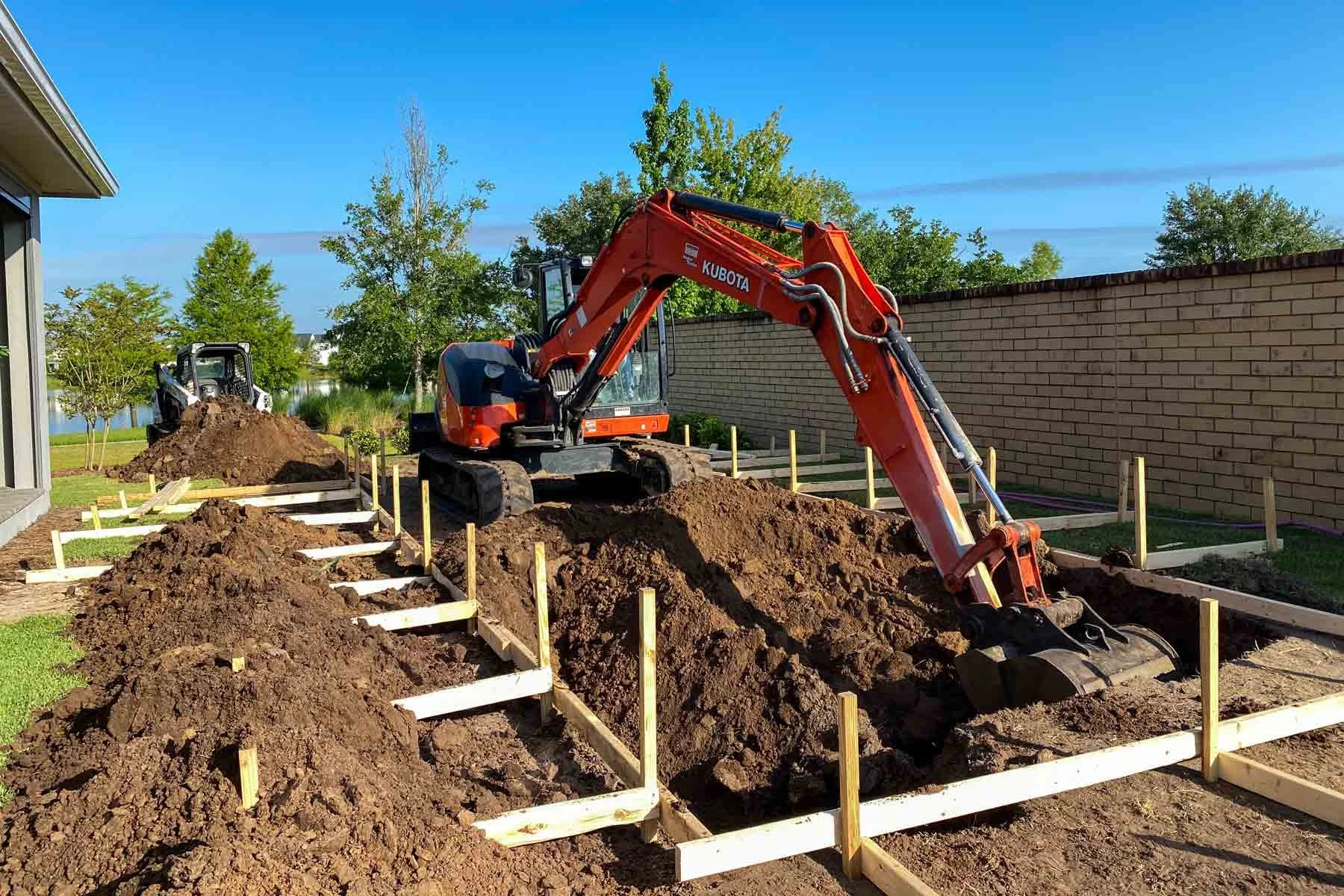 Construction site with three trenches being dug for a foundation, surrounded by wooden framing and piles of dirt. A small orange excavator is digging in the foreground, and another in the background, with a brick wall and trees under a clear blue sky.