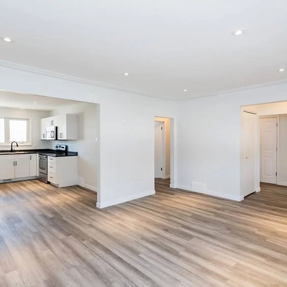 Empty living area with white walls, wood flooring, and a kitchen with white cabinets and black countertops.