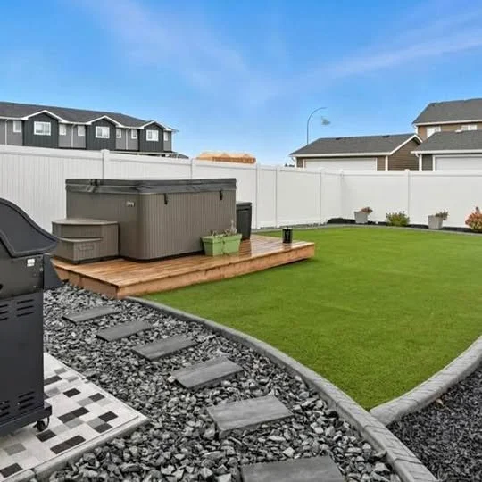 Backyard with green lawn, wooden deck, hot tub, small green storage container, and white privacy fence, with neighboring houses and blue sky in the background.