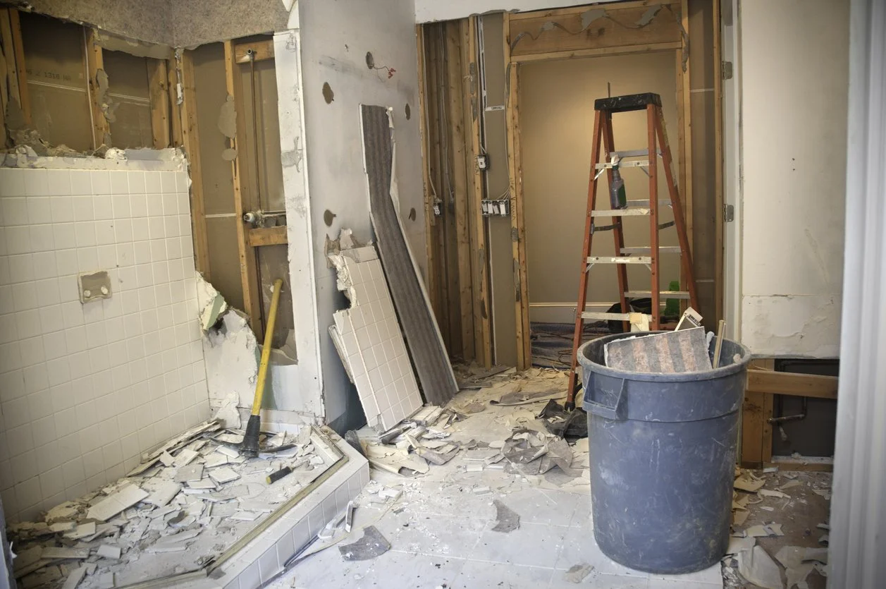 Bathroom under renovation with a damaged shower area, exposed wooden framing, a red ladder, a black cleaning spray bottle, and construction debris including broken tiles and drywall on the floor.