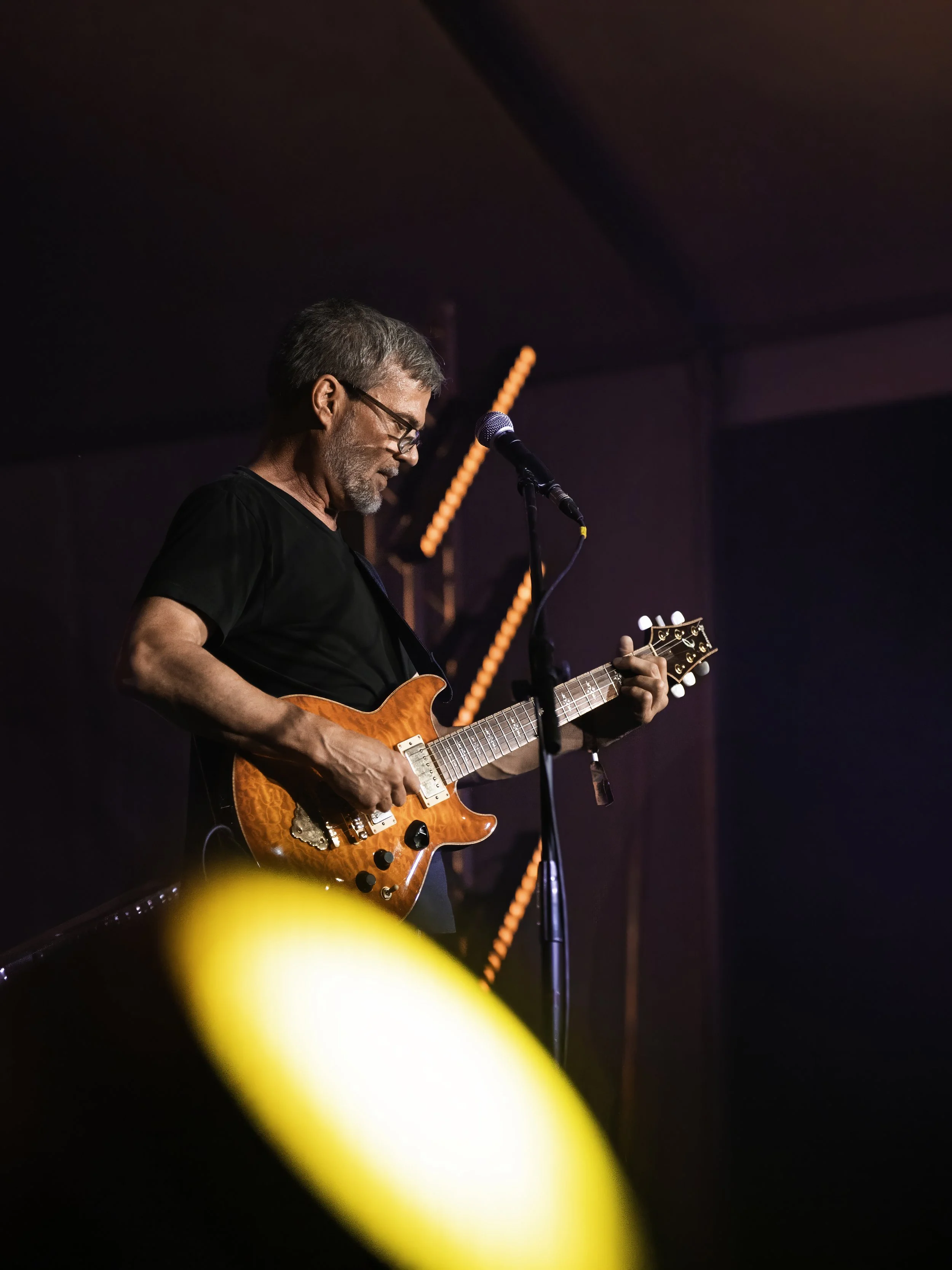 A man with glasses and gray hair playing an orange electric guitar on stage, with a microphone in front of him and stage lighting in the background.