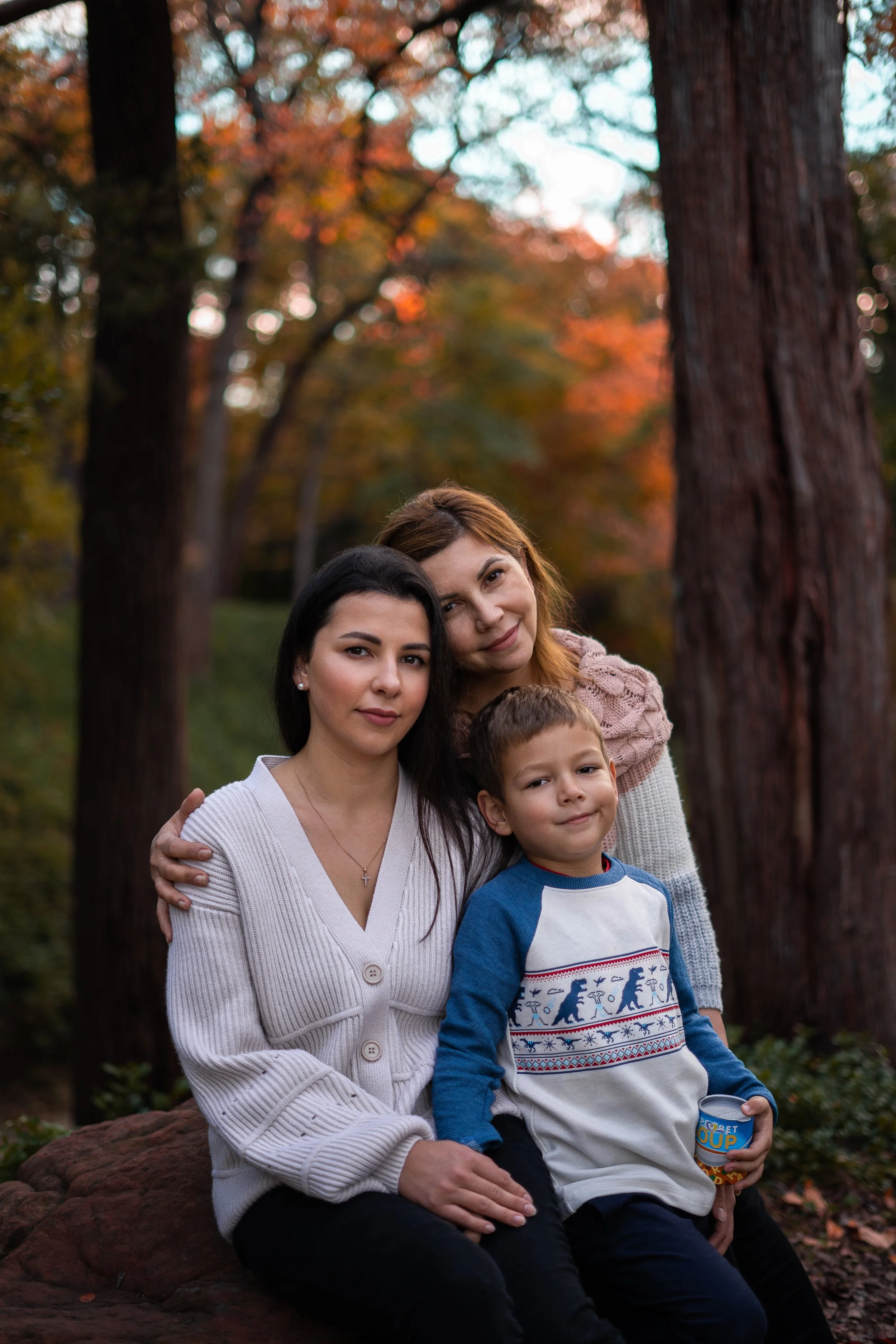 A group of four people, including two women and two children, sitting outdoors on a large rock with trees and autumn leaves in the background.