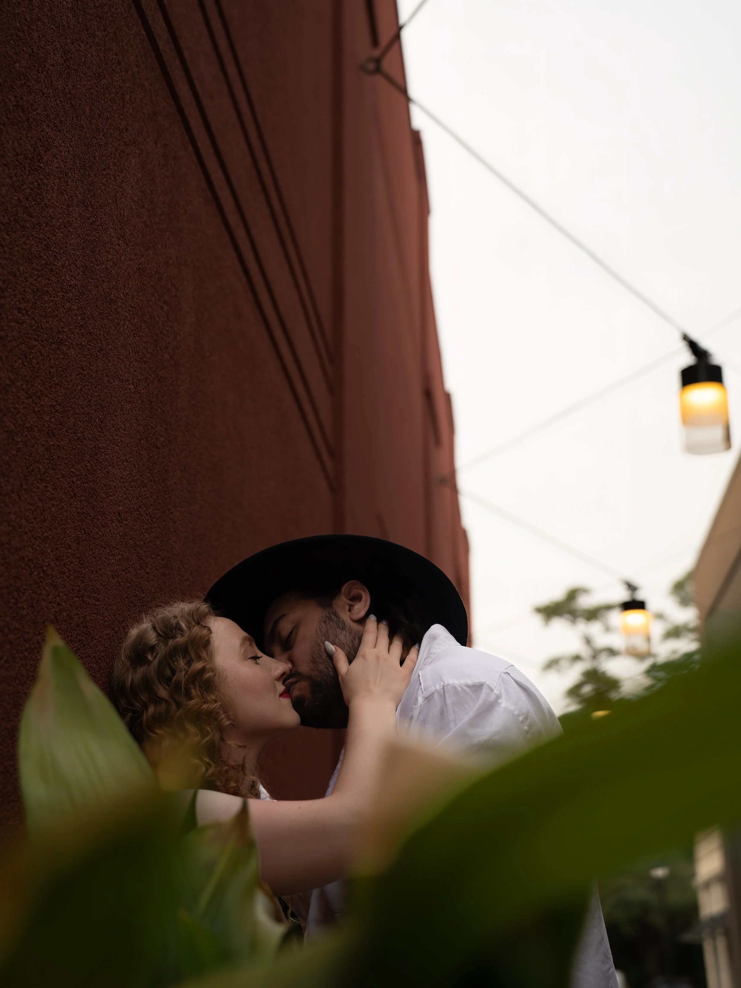 A romantic moment between a couple with their faces close, appearing to be about to kiss, surrounded by green plants and against an orange textured wall with outdoor lights.