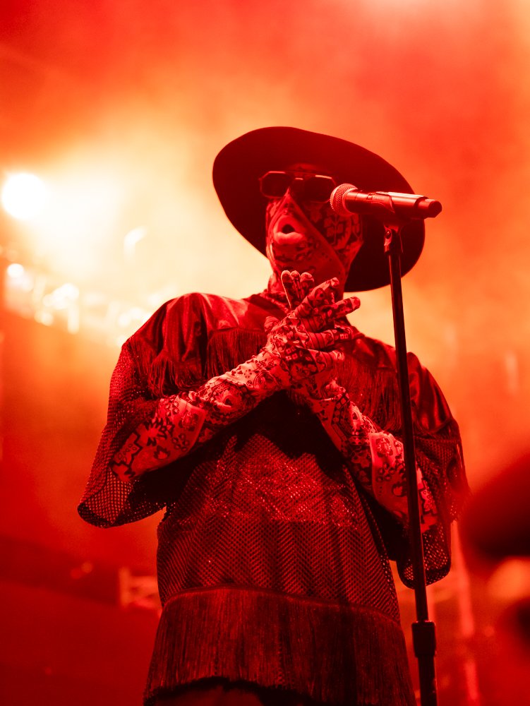 A person wearing a wide-brimmed hat, sunglasses, and a patterned face mask stands on stage with a microphone, under red stage lighting.
