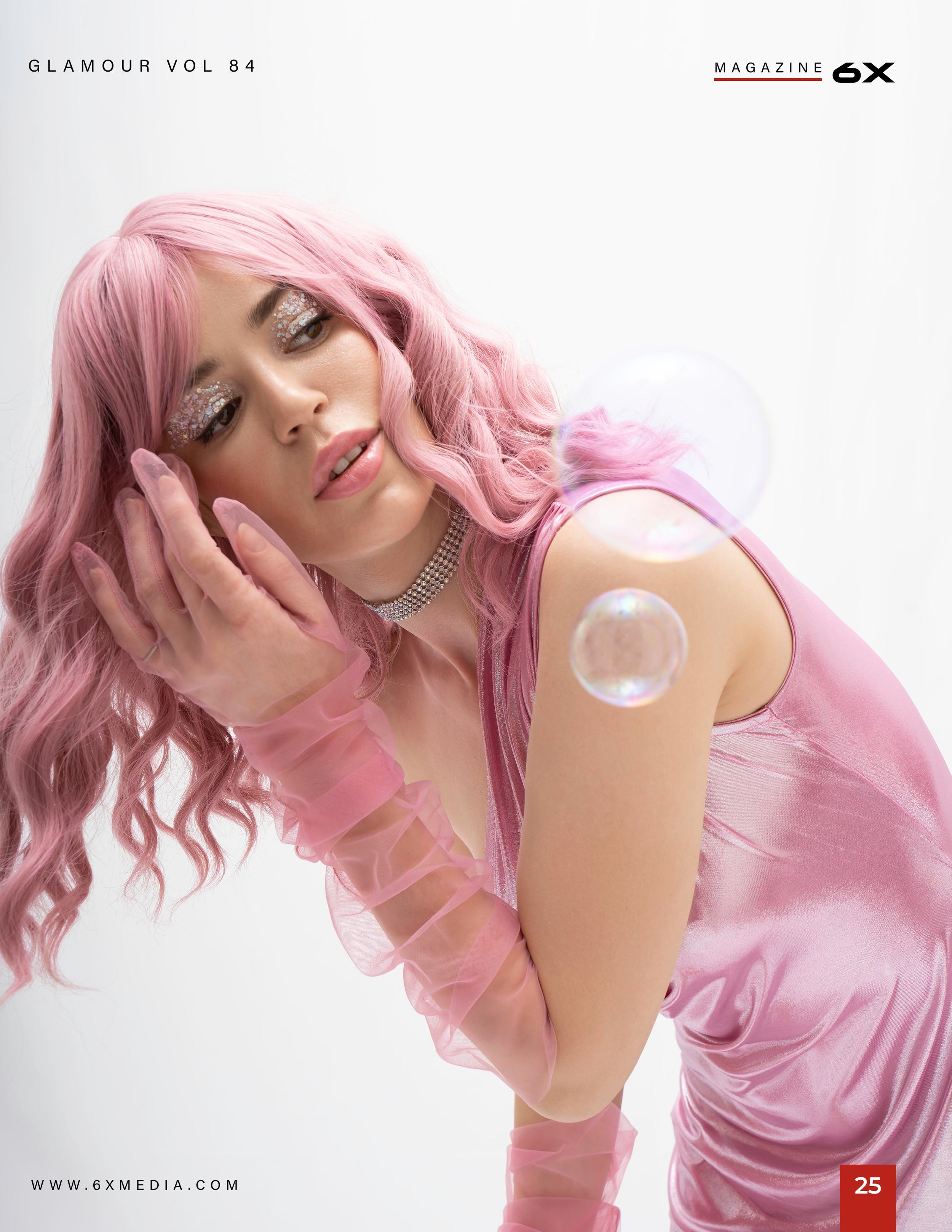 A woman with long pink hair and glittery makeup posing against a white background, wearing pink clothing and translucent pink gloves.