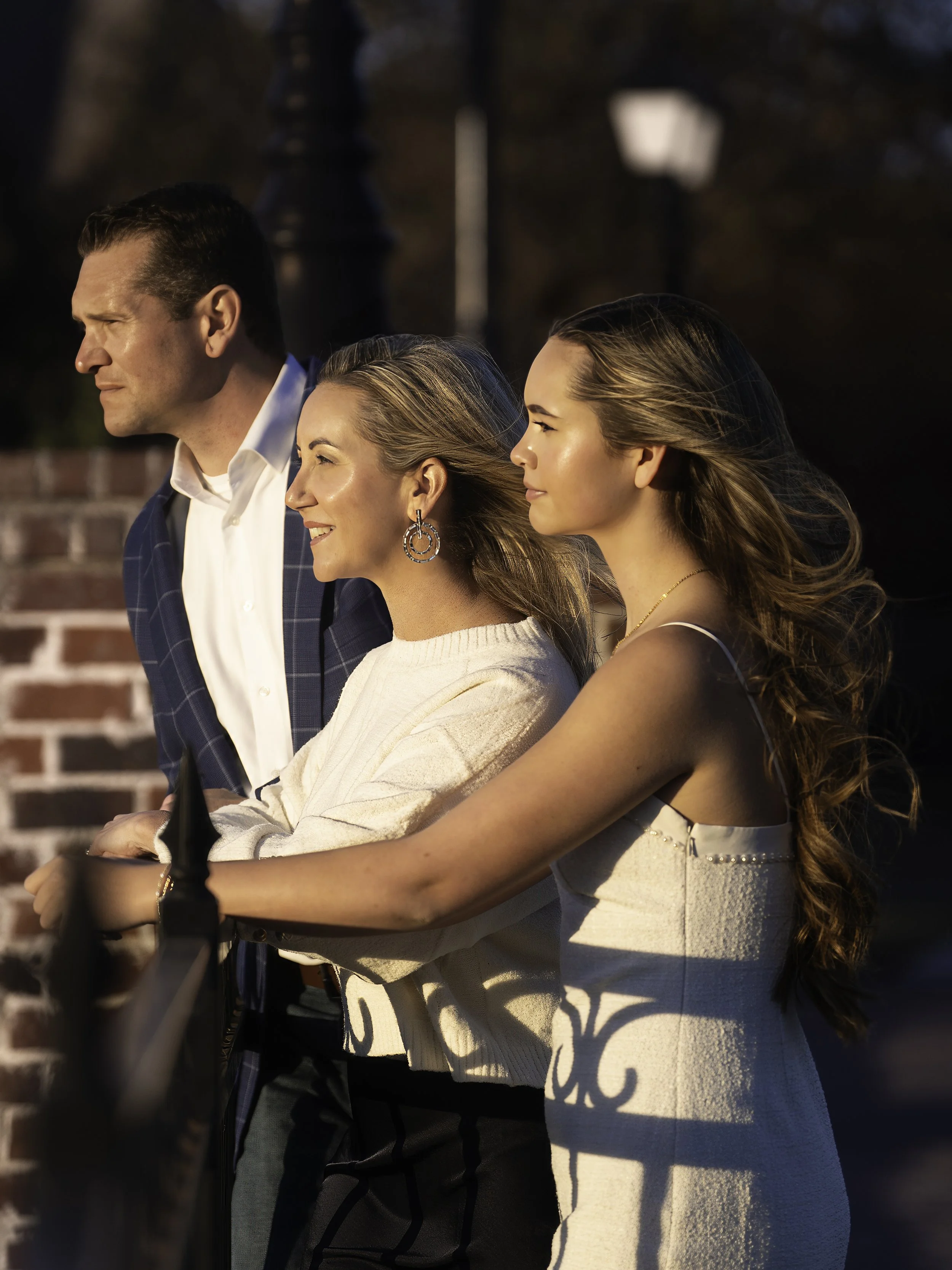 Three people, two women and one man, standing outdoors in the sunlight. They are dressed formally, with the woman in a white dress, the man in a blue suit, and the other woman in a light-colored top. They are gazing in the same direction, with a brick wall and a lamp post visible in the background.