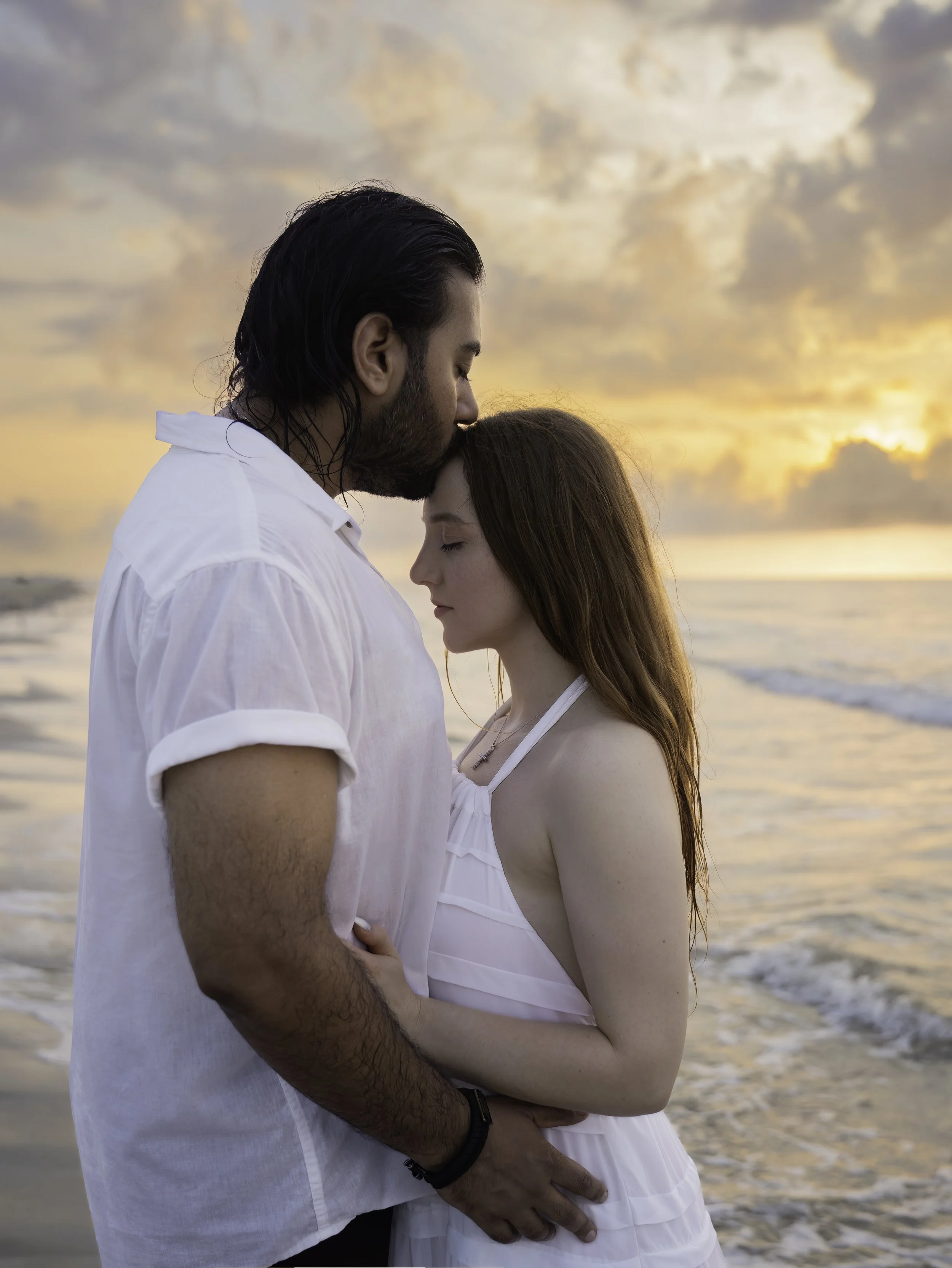 A man and woman standing close together on the beach during sunset, with the man kissing the woman's forehead, both with eyes closed.