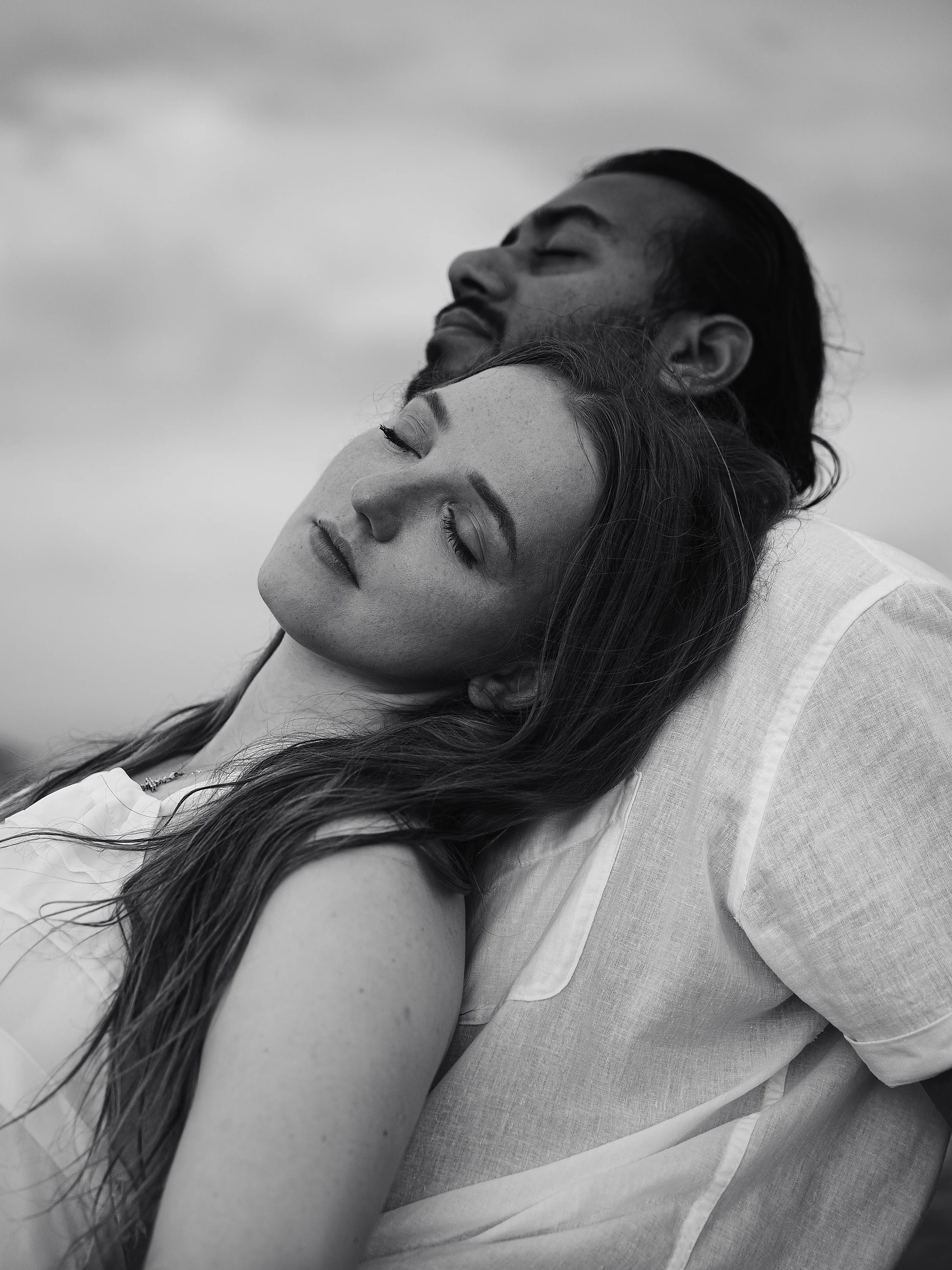 Black and white photograph of a woman with long hair, lying with her eyes closed, and a man with a beard resting his head against her forehead with his eyes closed. The scene suggests an intimate and peaceful moment.