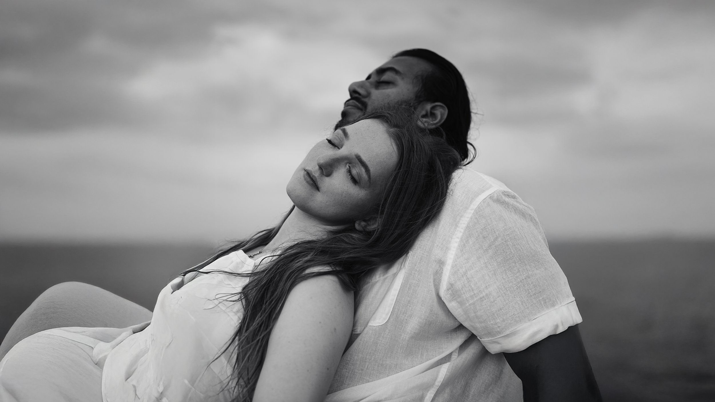 A black and white photo of a man and woman with closed eyes, leaning on each other against a cloudy sky, conveying tranquility and intimacy.