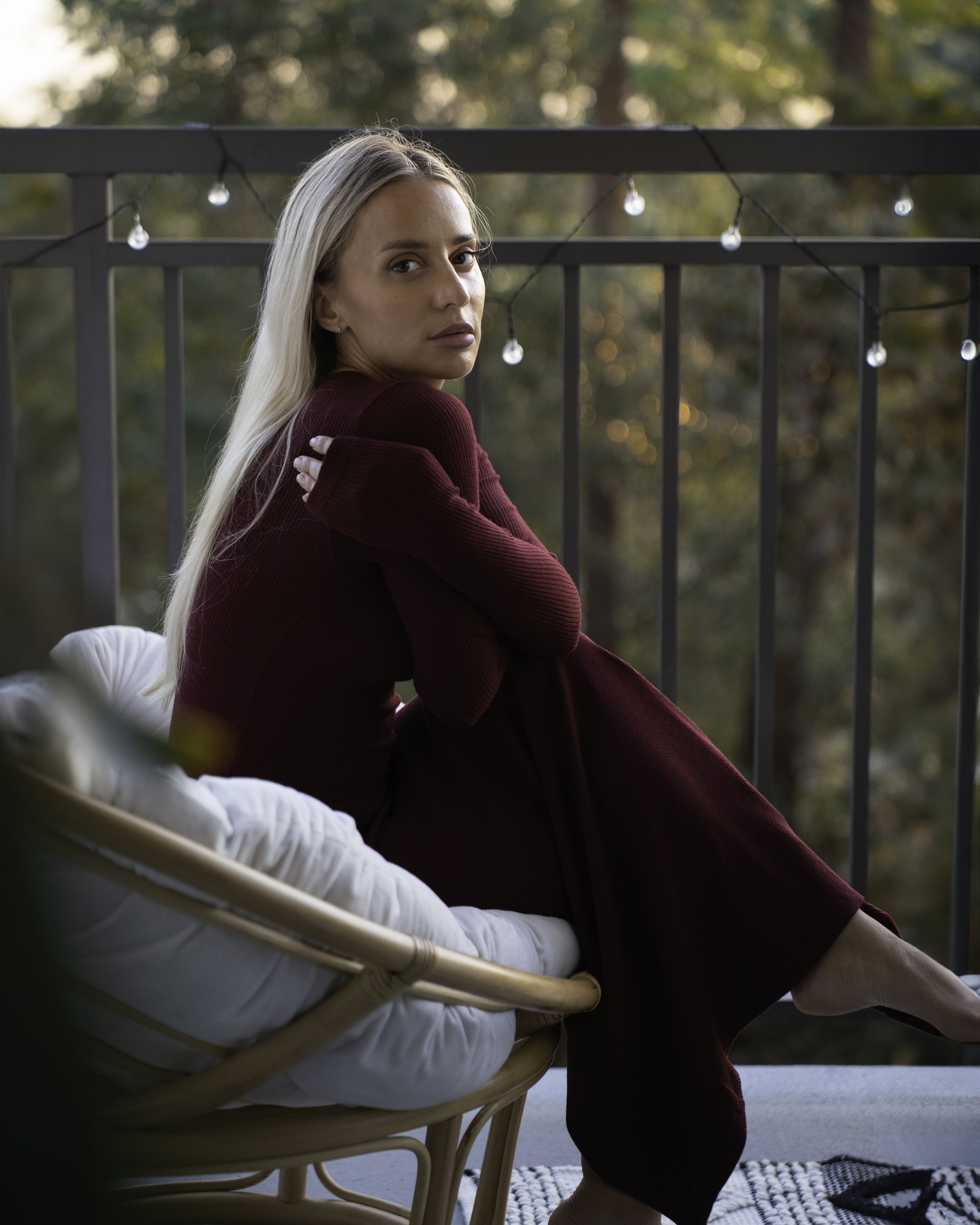 A woman with long blonde hair sitting outdoors on a balcony railing, looking over her shoulder at the camera, with string lights hanging in the background and trees behind her.