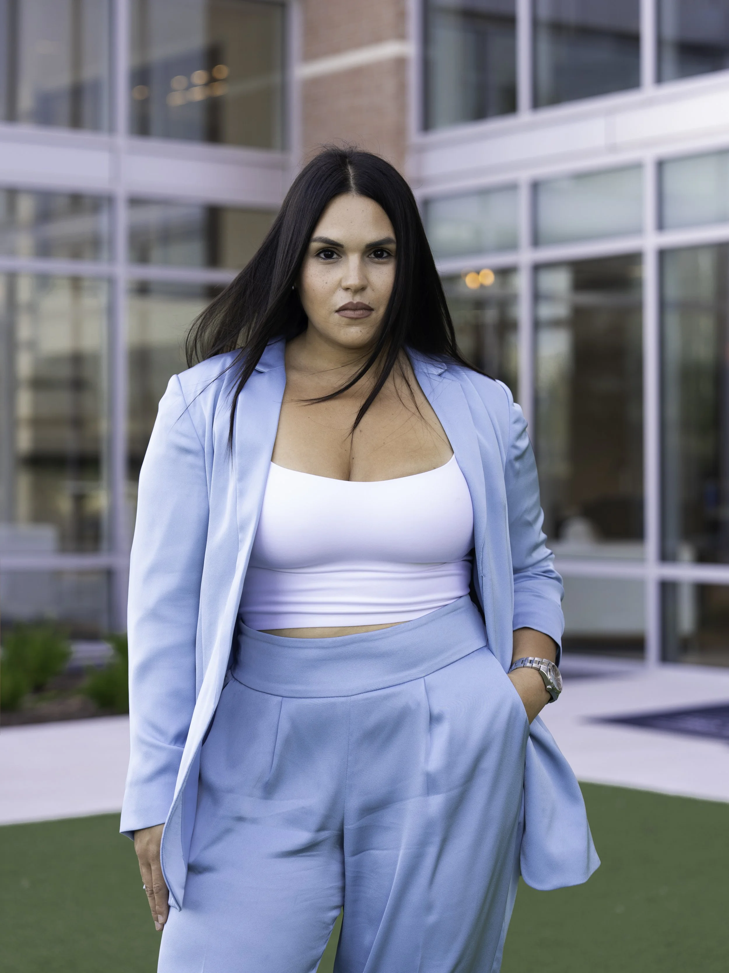 A woman with long black hair wearing a light blue suit and silver watch, standing outside in front of a modern glass building.