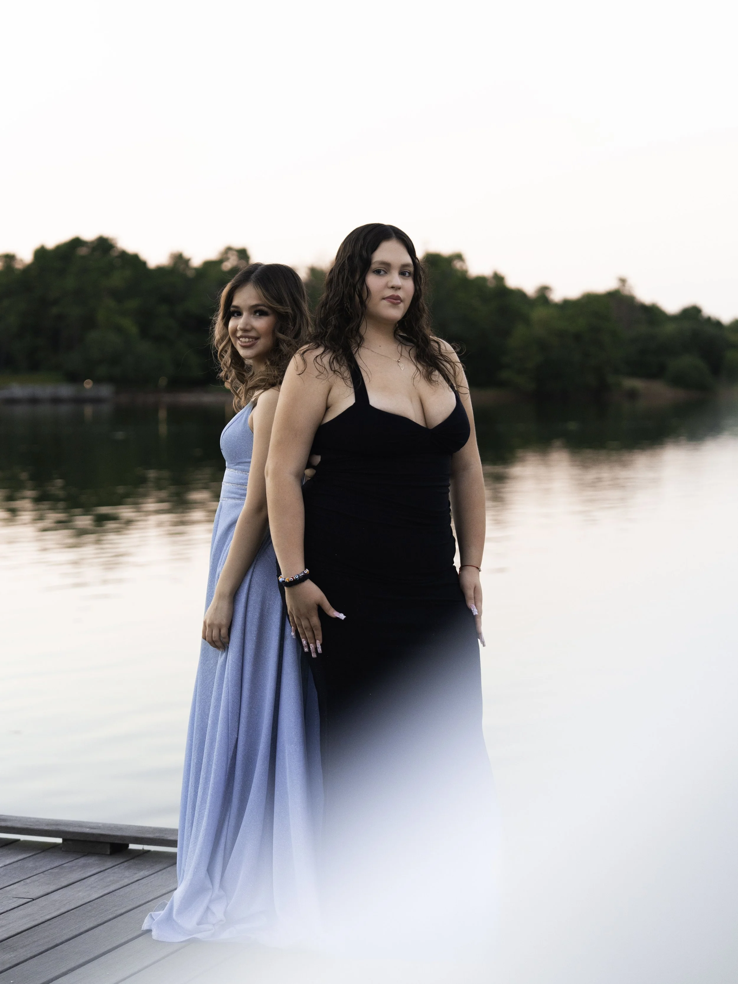 Two young women in formal dresses standing on a dock near a lake, with trees in the background during sunset.