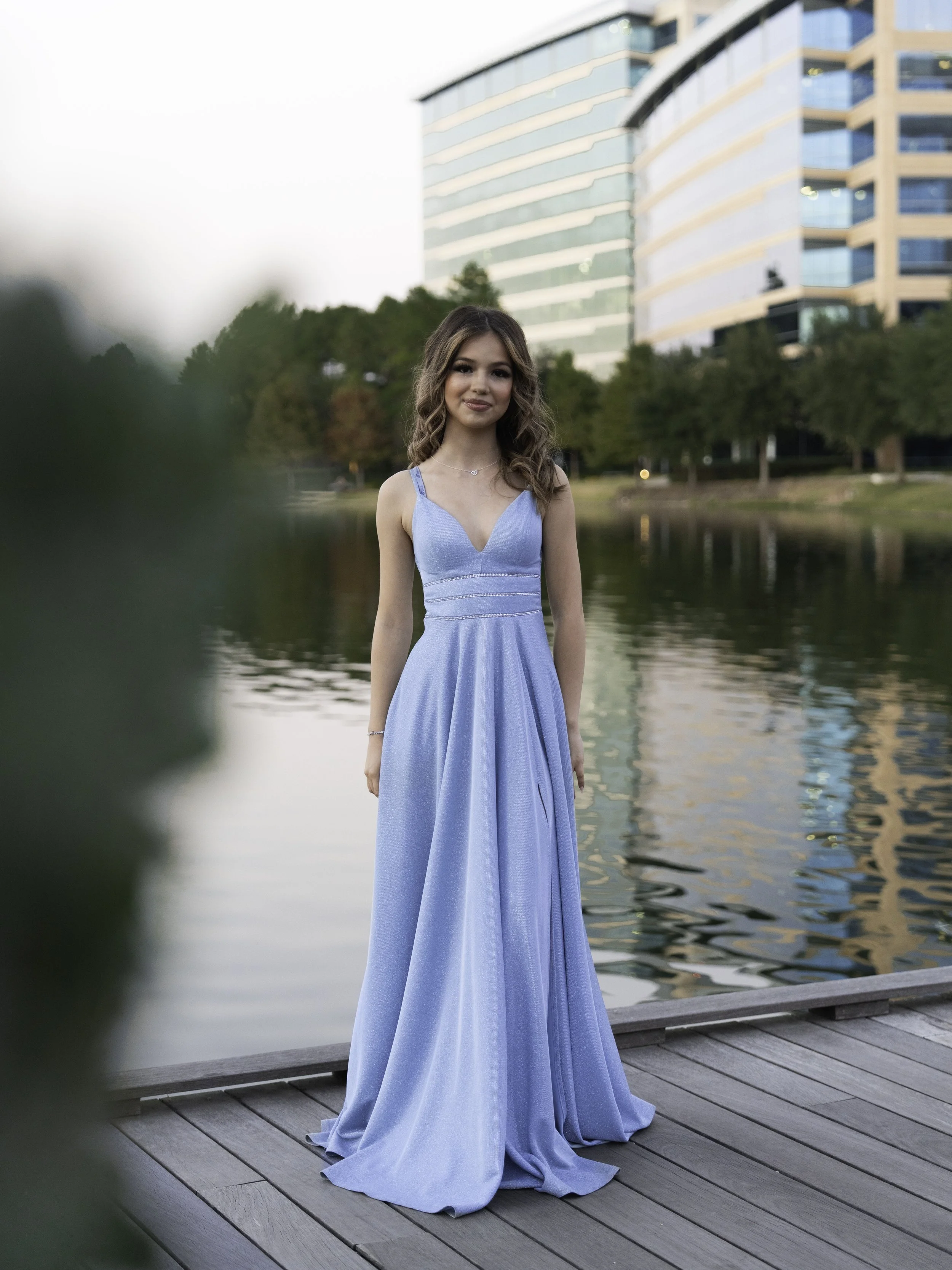 A young woman in a strapless, light blue gown standing on a wooden dock beside a body of water, with modern glass buildings and trees in the background.