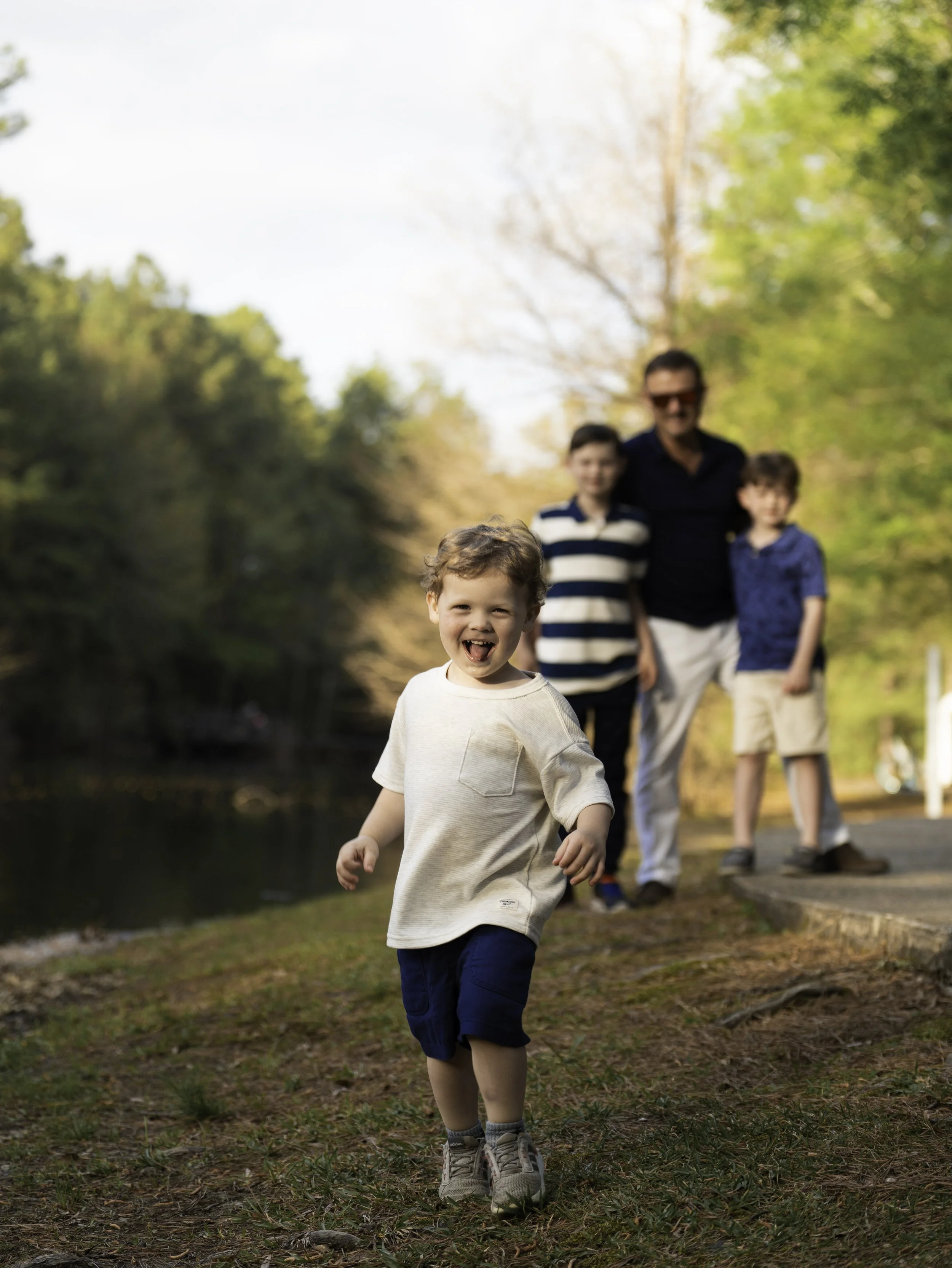 A young boy in a white shirt and blue shorts running on a nature trail, with a family of three in the background—an adult male and two children, all walking on the trail surrounded by trees with green and autumnal foliage, during what appears to be late afternoon.
