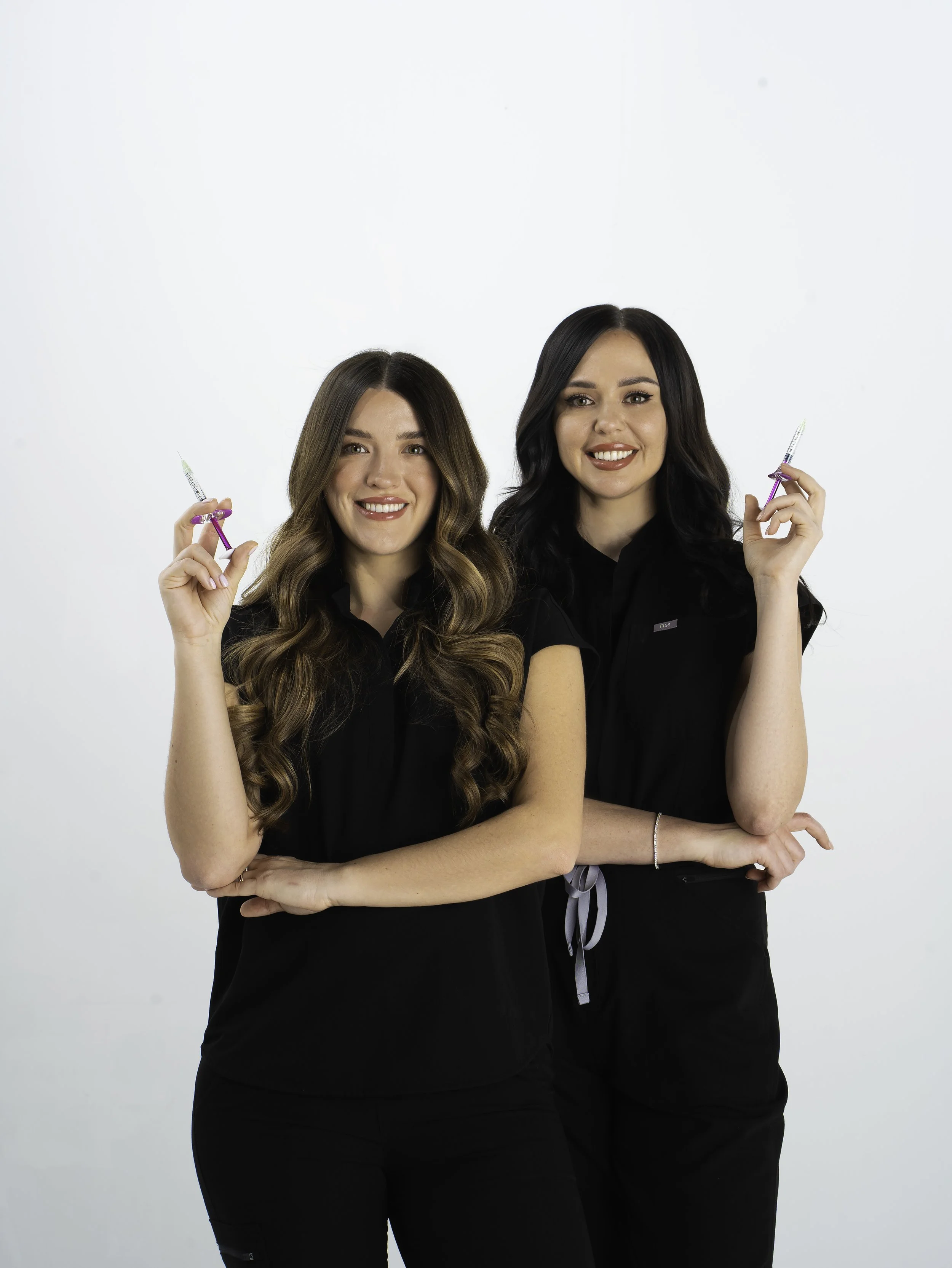 Two smiling women with long hair, wearing black tops, holding syringes, posing against a plain white background.