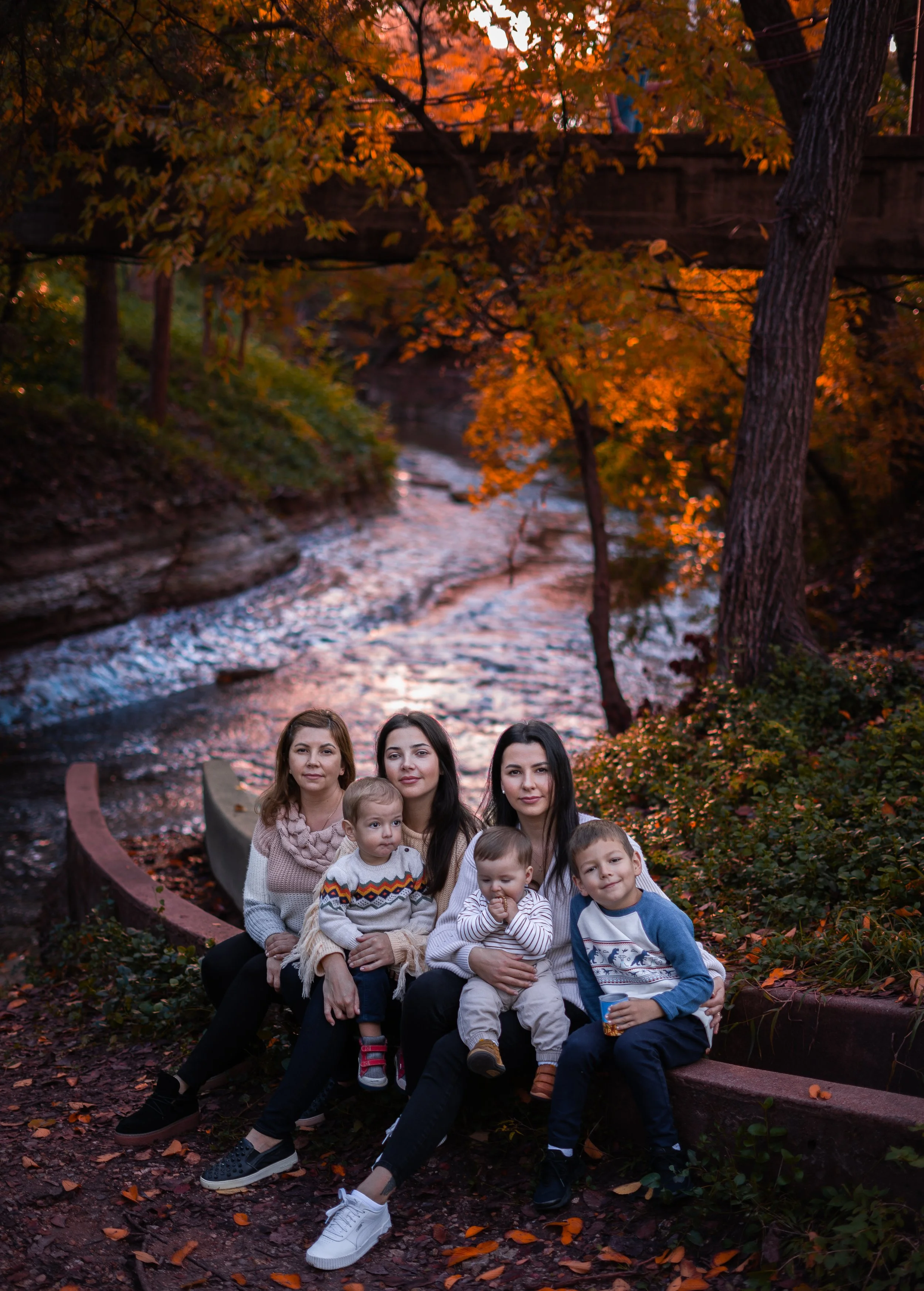 A family of seven sitting on a curved bench outdoors during autumn, with a river and trees with orange and yellow leaves in the background. The sky is partly cloudy, and the scene has warm, natural lighting.