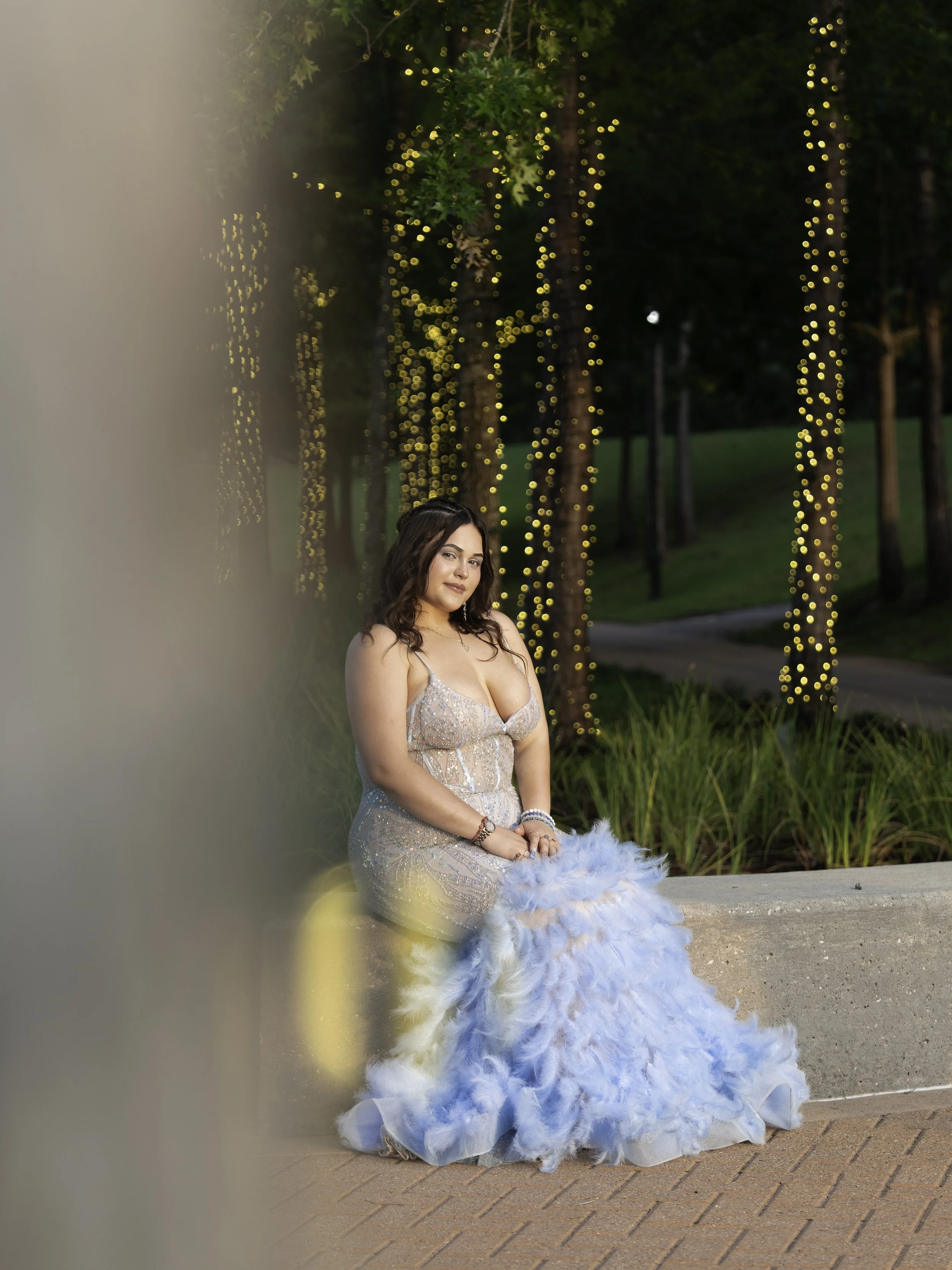 A woman in an elegant, sparkly evening gown with a large blue feathered train, sitting outdoors at dusk near trees with string lights.