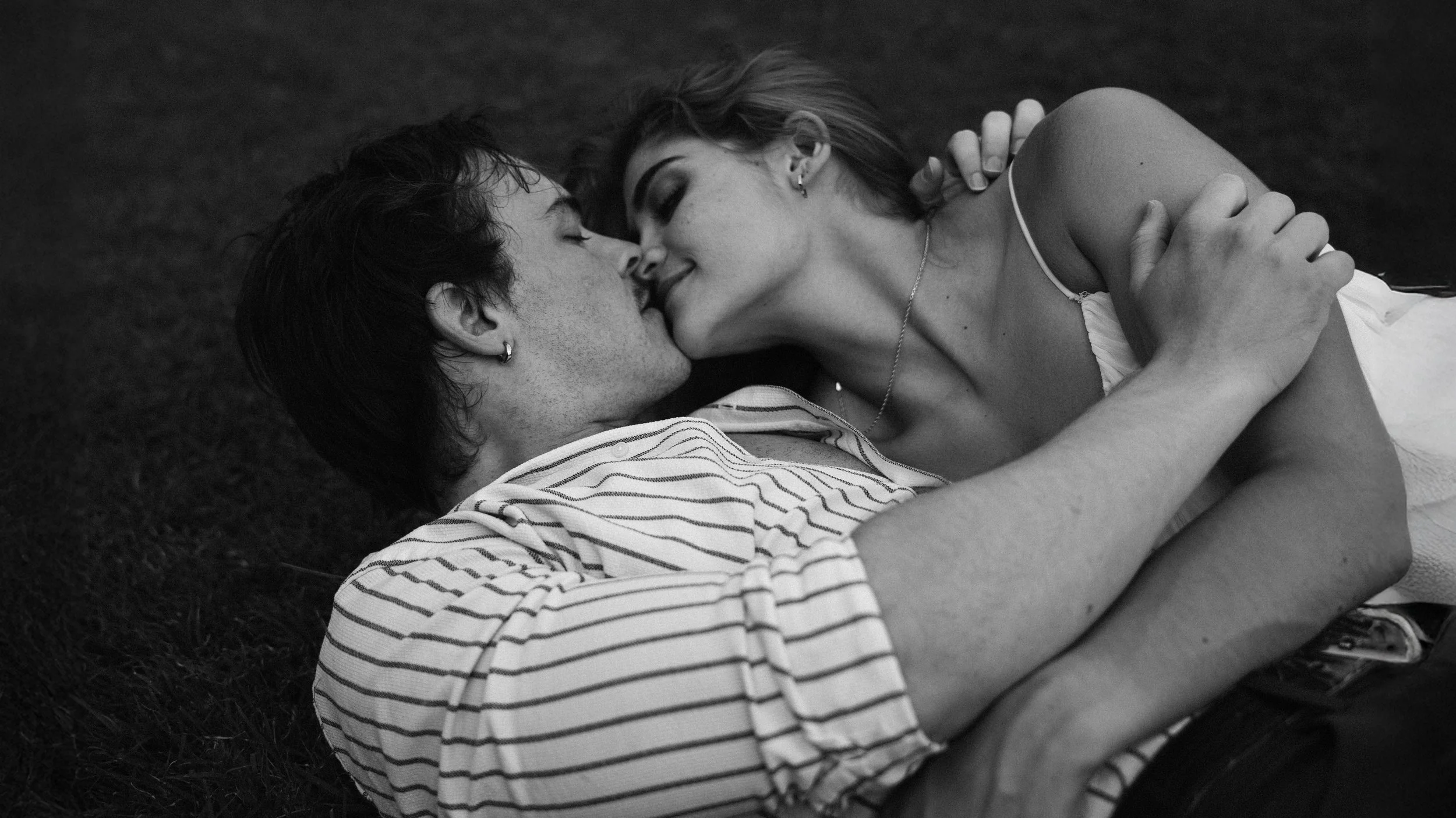 A black and white photo of a couple lying on the grass, touching their foreheads together, with their eyes closed, sharing an intimate moment.