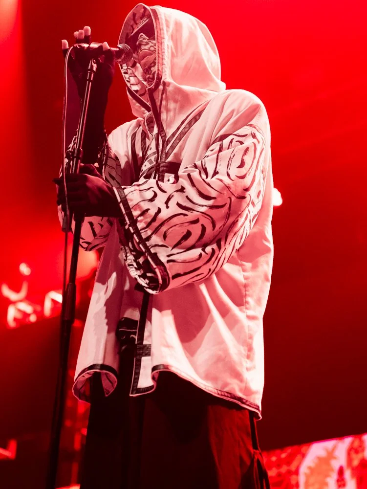 Person wearing a hoodie with a tiger stripe pattern, singing into a microphone on a stand, illuminated by red stage lighting.