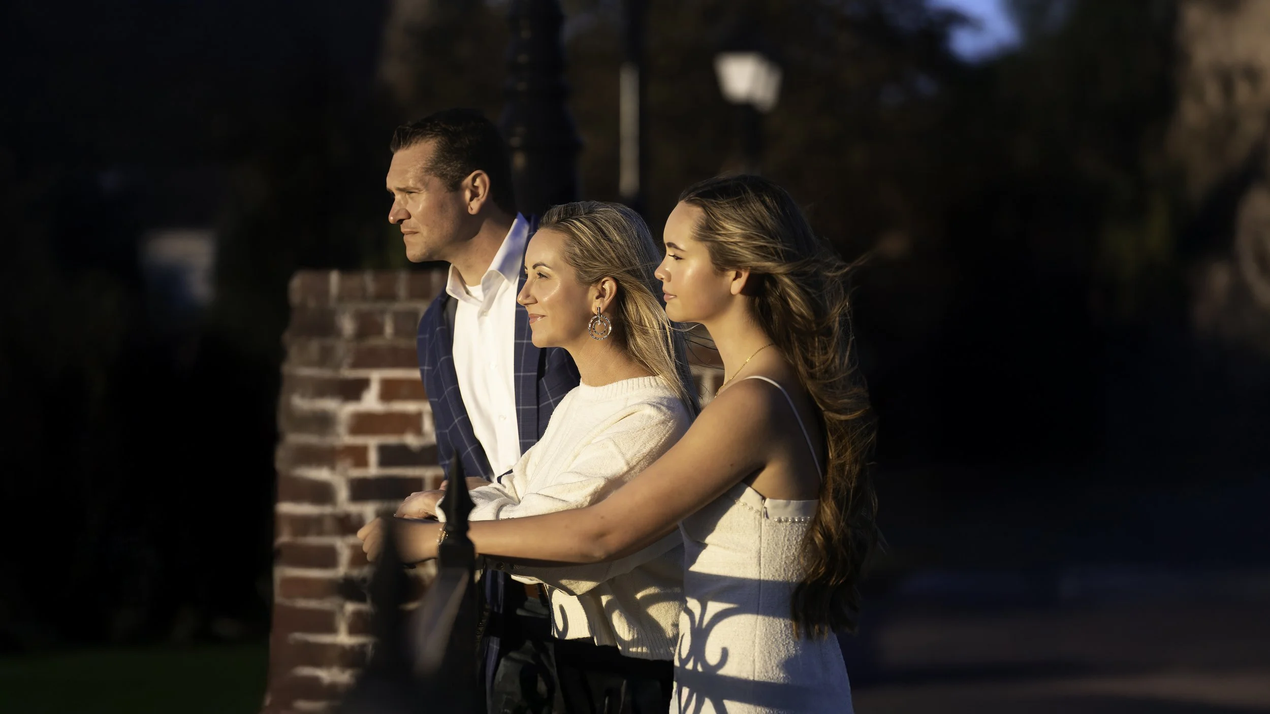 Three people, two women and one man, standing outdoors by a brick wall with a railing, during sunset, with trees and street lamps in the background.
