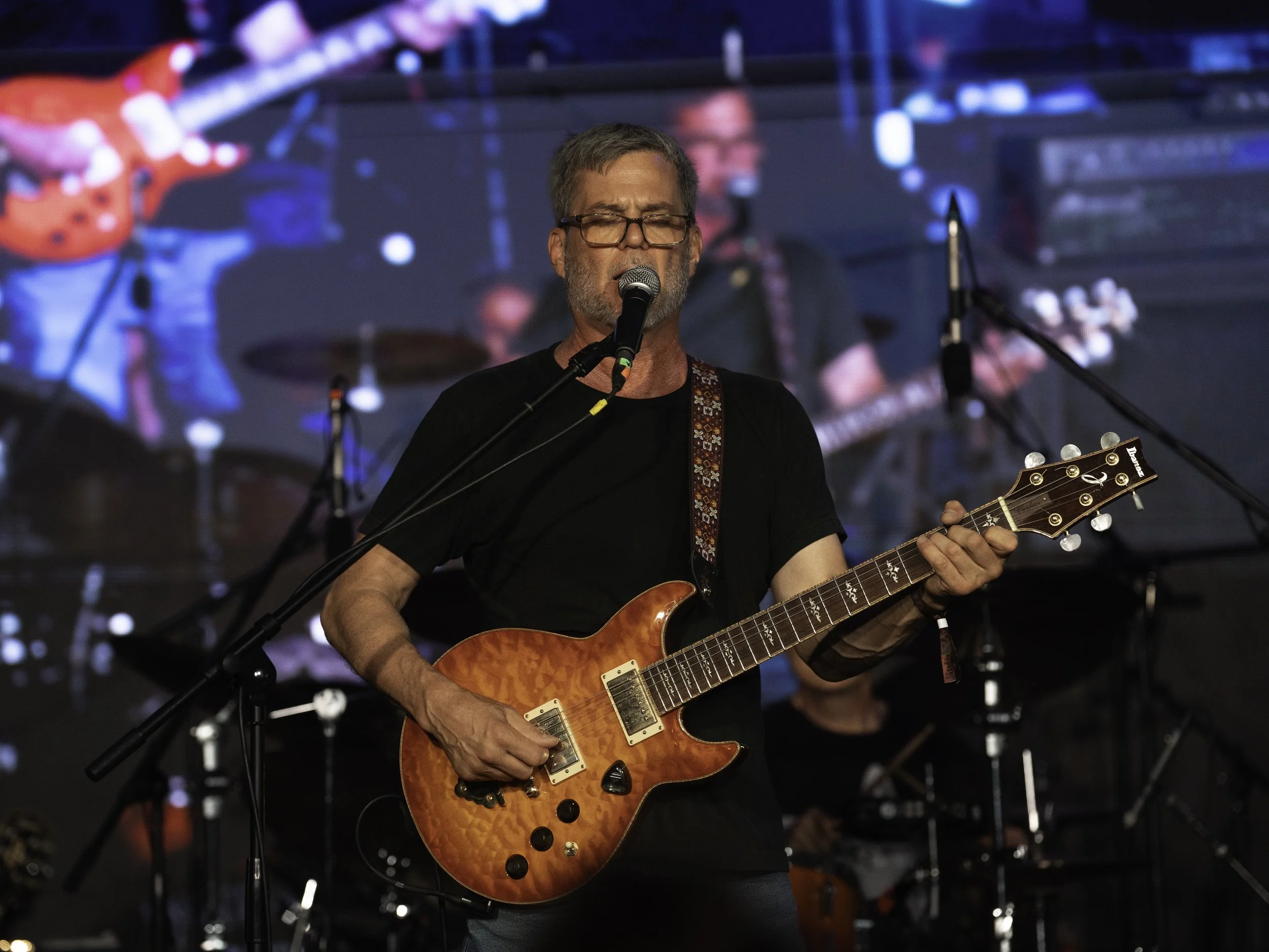 A man with glasses and gray hair singing into a microphone and playing an orange electric guitar on stage with blue and red lighting.
