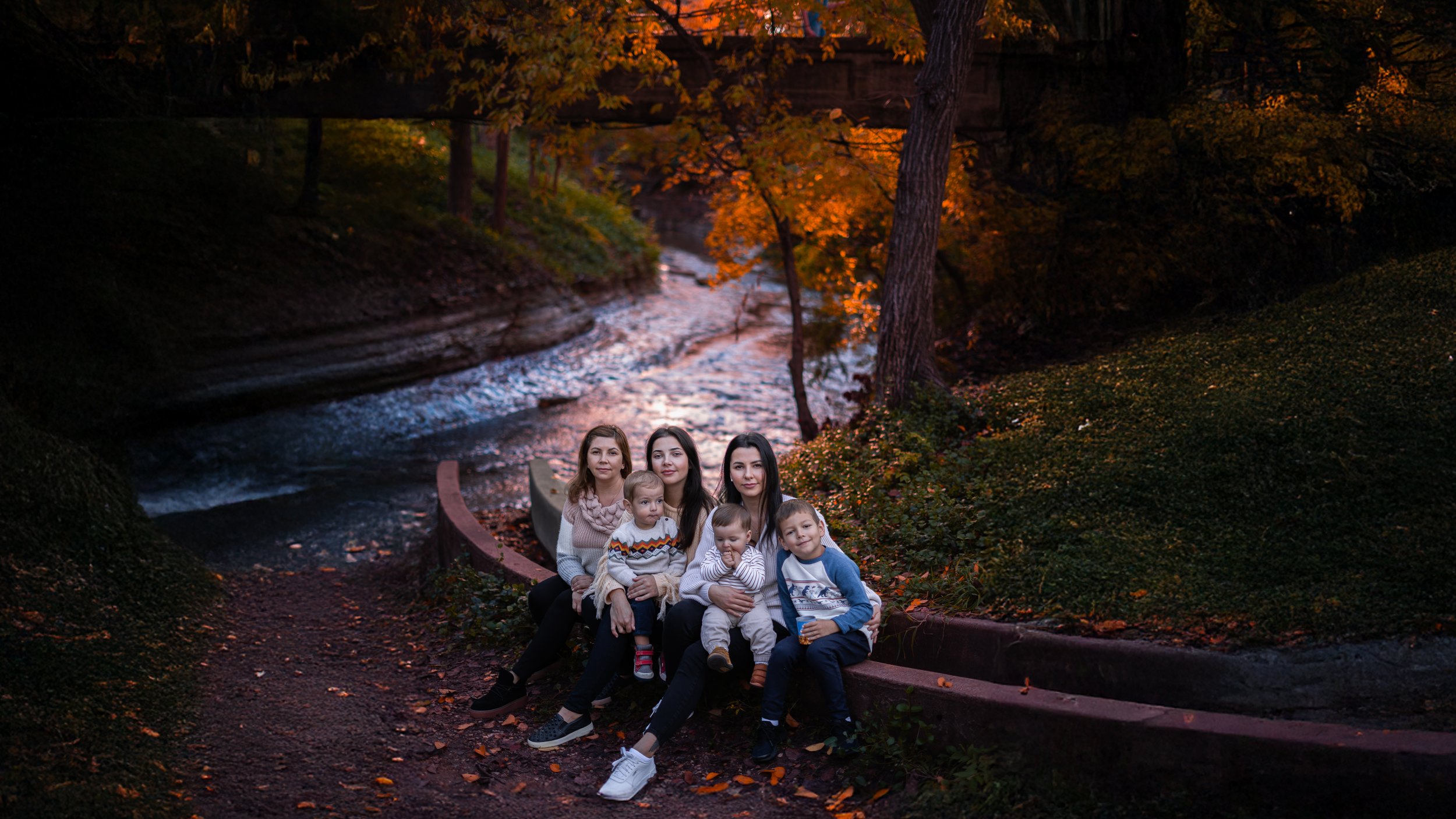 Group of five women and four children sitting together on a curved park bench next to a river with autumn foliage in the background.