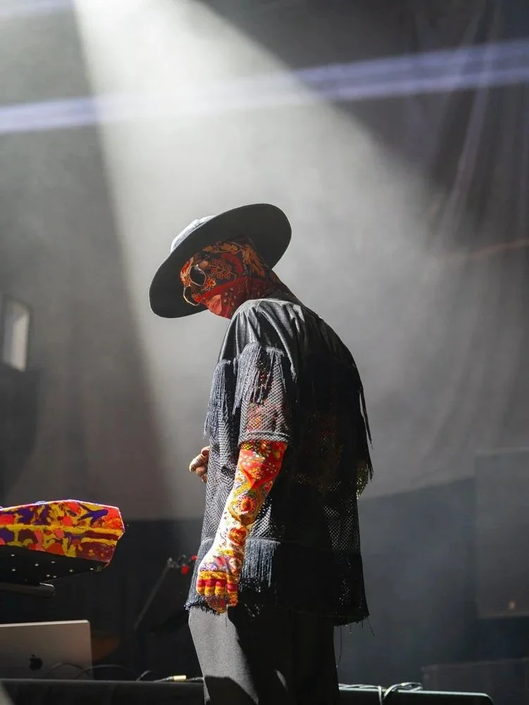 A person wearing a large black hat, a colorful face mask, a black mesh shirt, and colorful patterned long sleeves, standing on stage with bright stage lights shining down.