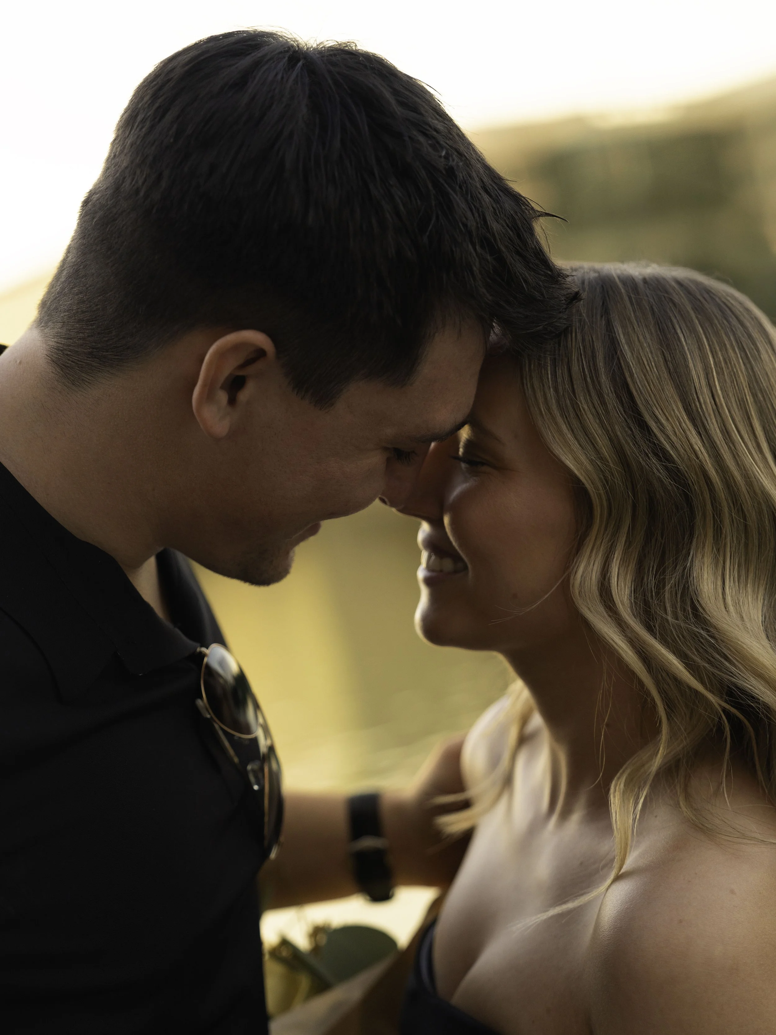 A close-up of a young couple with foreheads touching, smiling, outdoors during sunset, with blurred landscaping in the background.