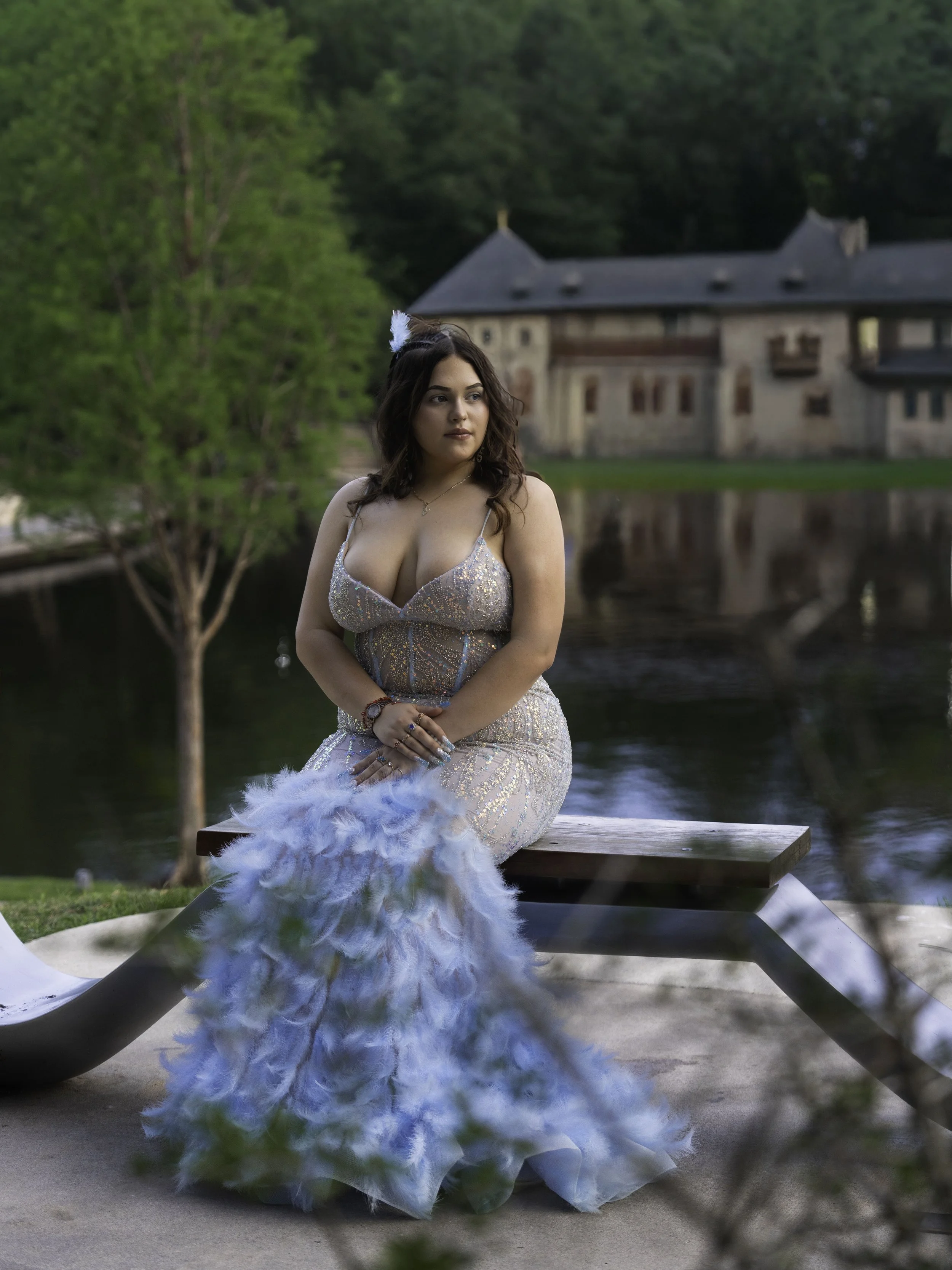A young woman in a sparkly dress with a feathered boa, sitting on a park bench near a body of water, with lush green trees and an old brick building in the background.