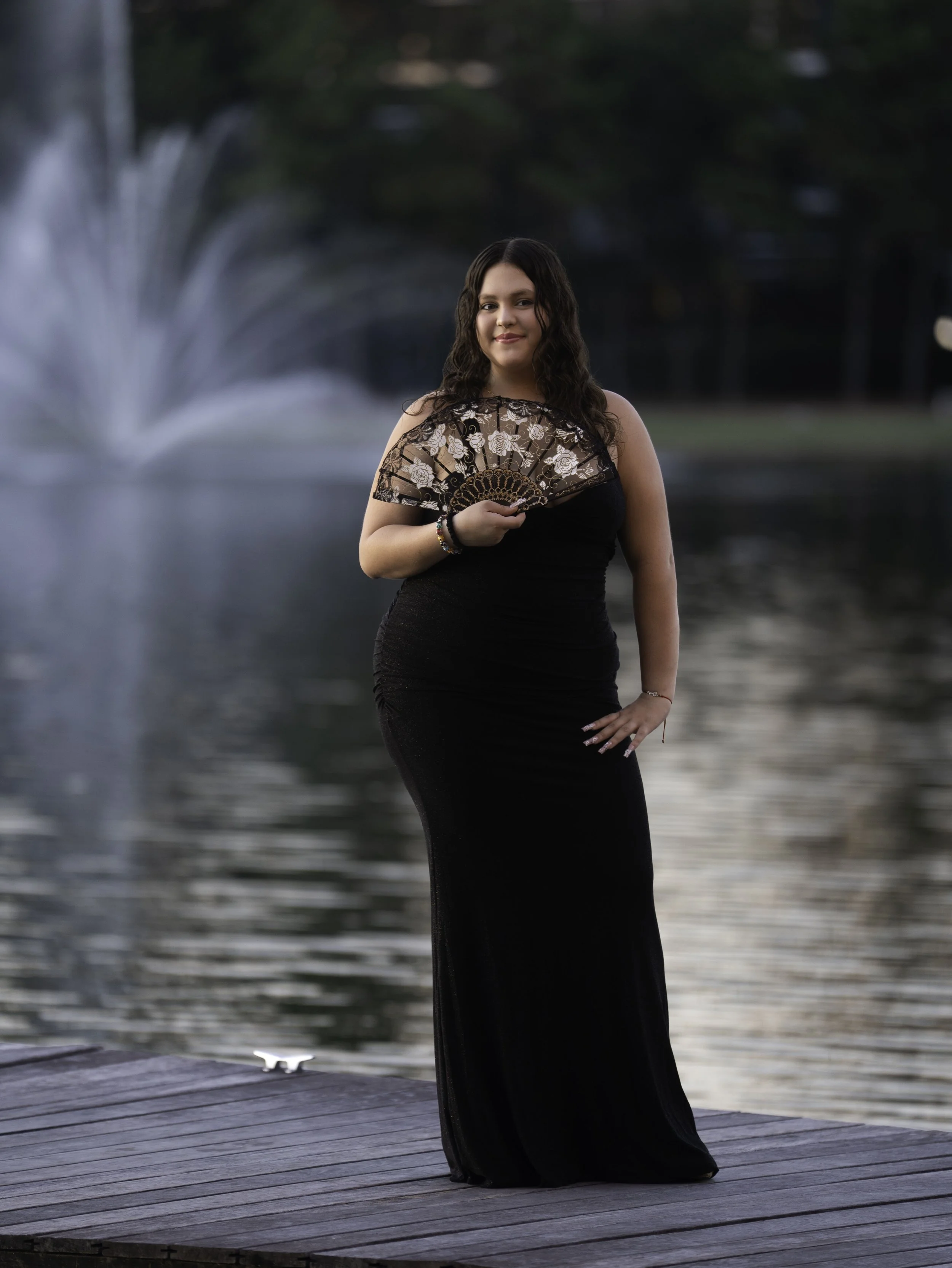 A woman in a long black dress holding a decorative fan stands on a wooden dock near a body of water with a fountain in the background. It's likely evening or early morning.