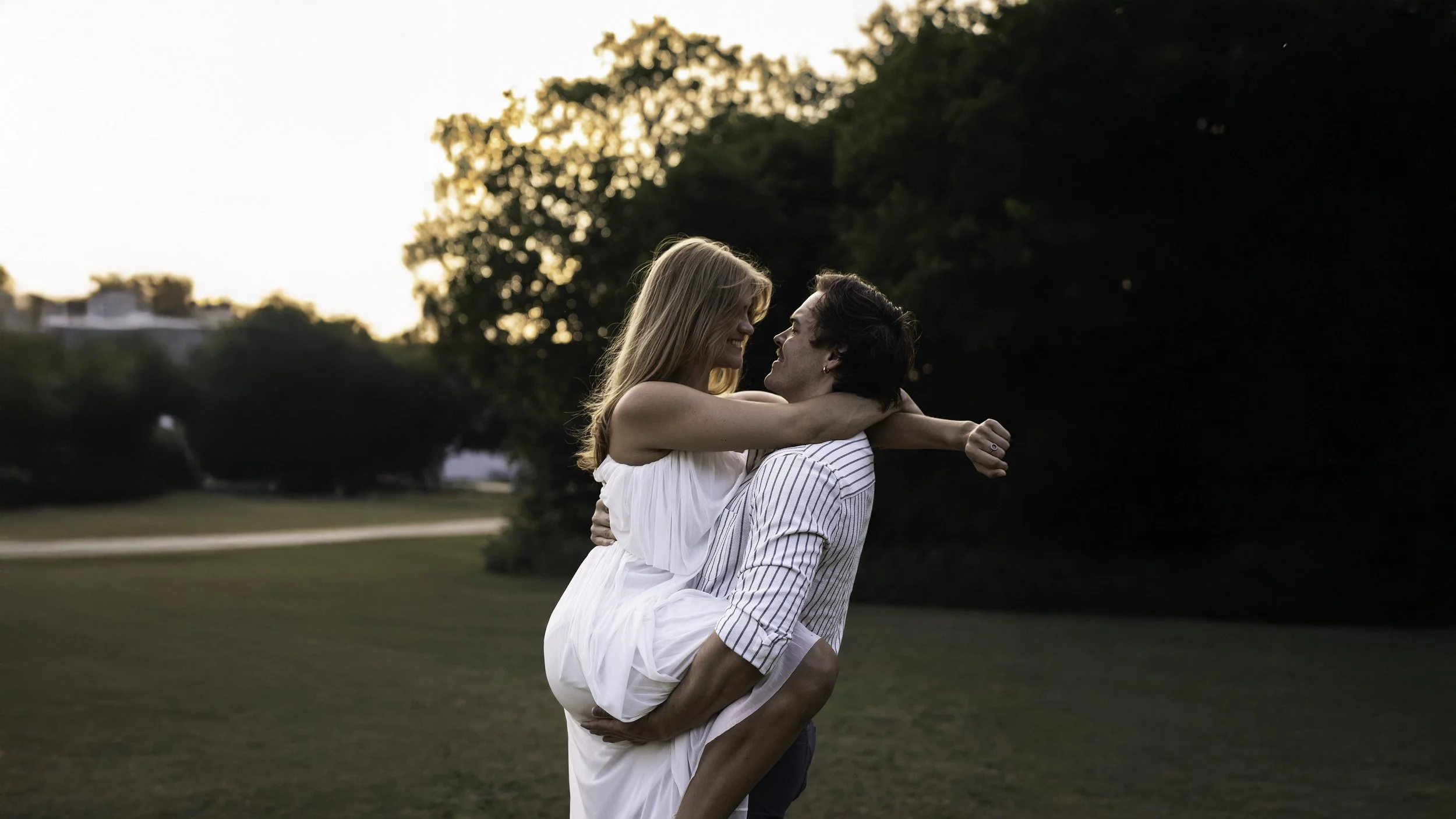 A couple outdoors at sunset; the woman, with blonde hair, is smiling and embracing the man sitting on the grass, with his arms around her waist, surrounded by trees.