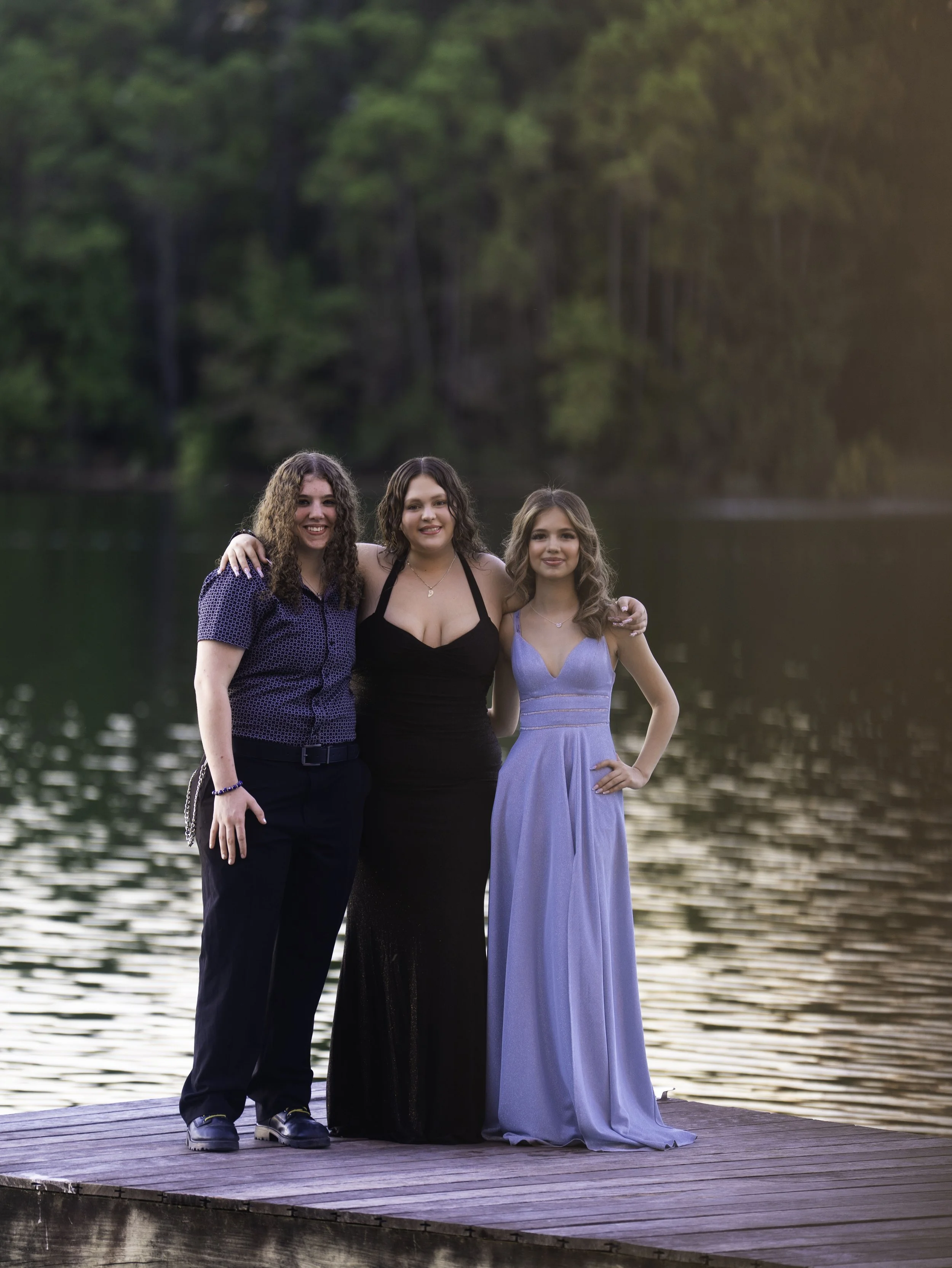 Three women standing on a wooden dock by a lake with trees in the background, smiling at the camera at sunset.