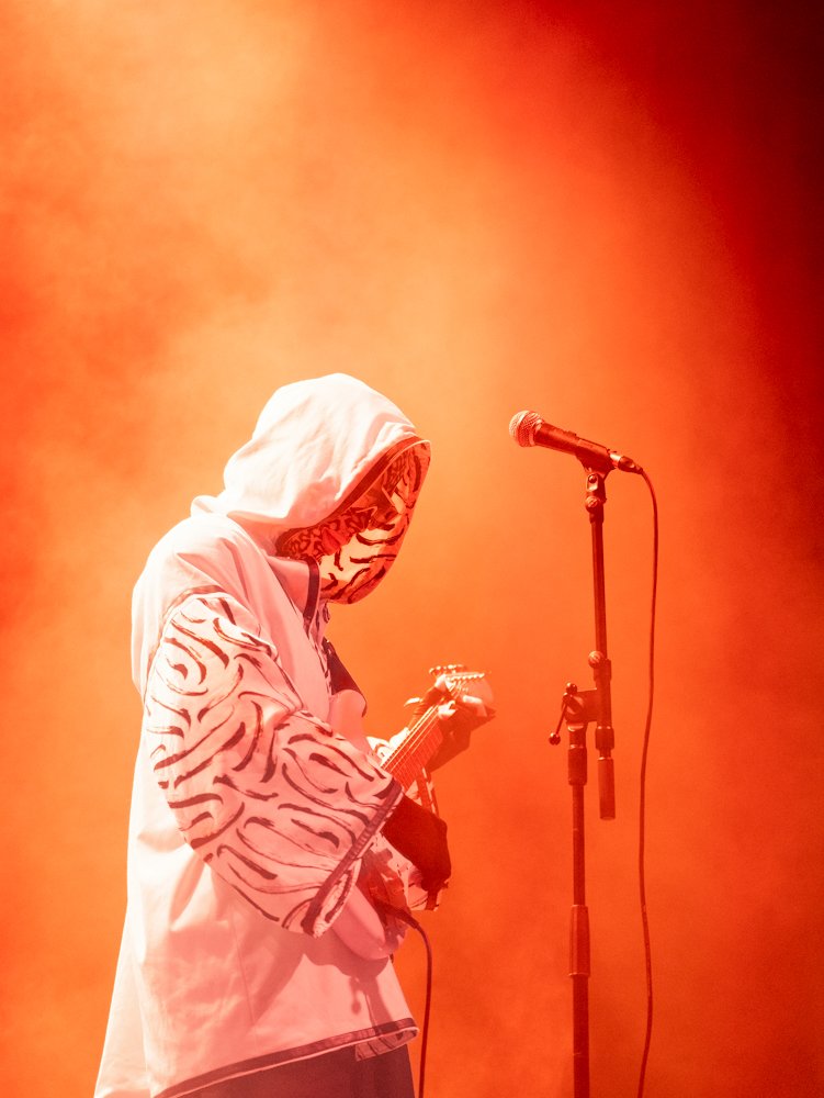 A person wearing a hoodie with tiger print on the sleeves playing an electric guitar on stage with orange lighting and a microphone stand.
