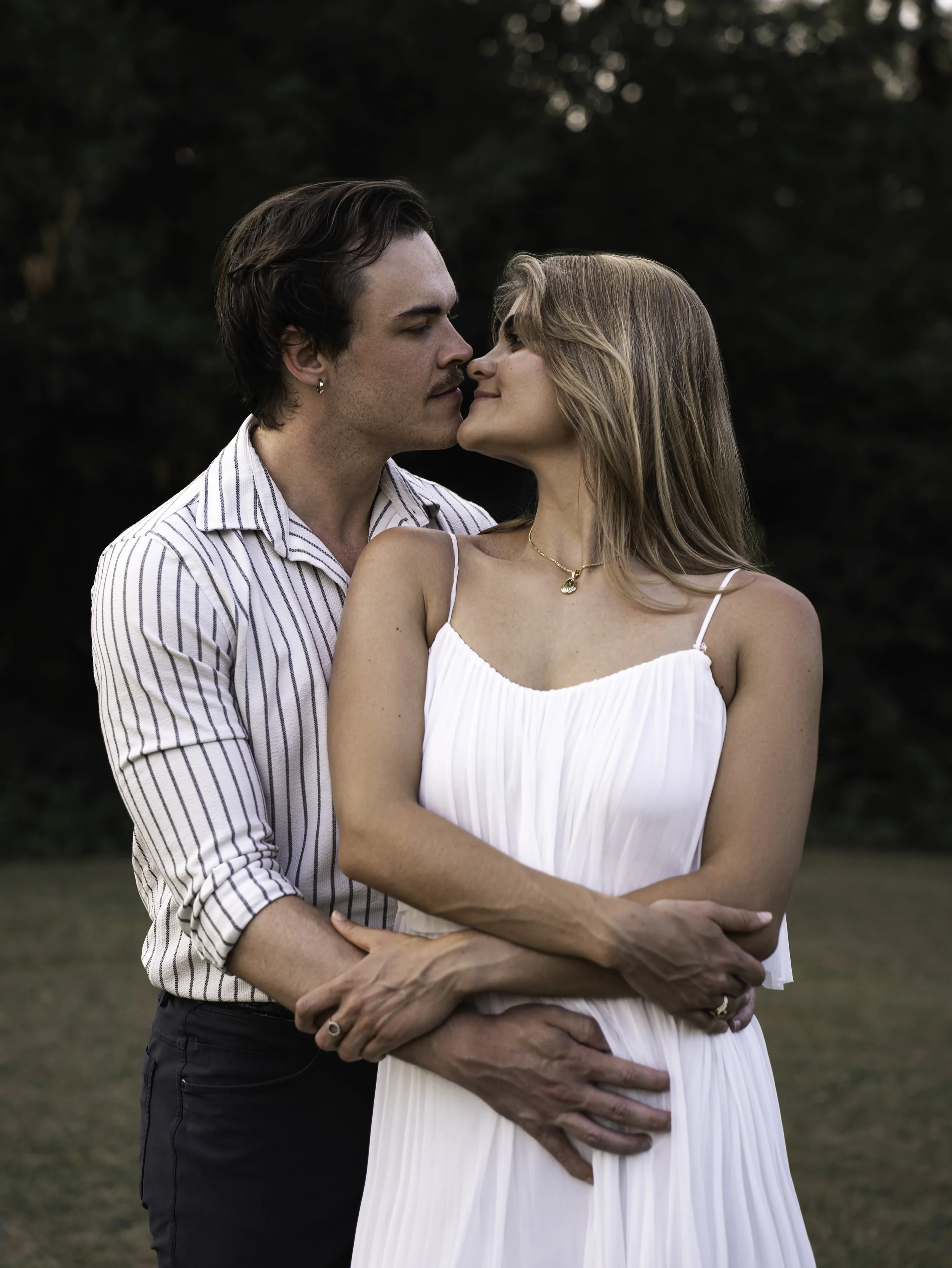 A young man with dark hair, earrings, and a striped shirt, standing closely with a young woman with blonde hair, wearing a white dress and jewelry, outdoors with trees in the background, engaged in an intimate moment.