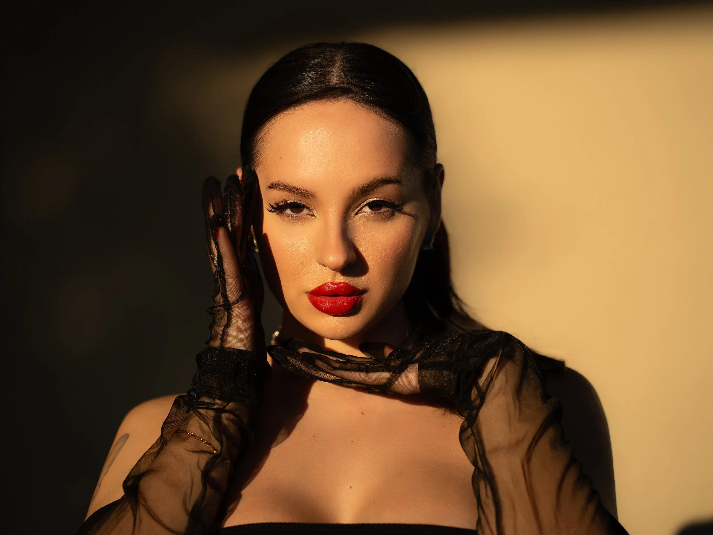 A woman with dark hair and red lipstick poses with her hand near her face, wearing black lace gloves, against a gradient background from dark to light.