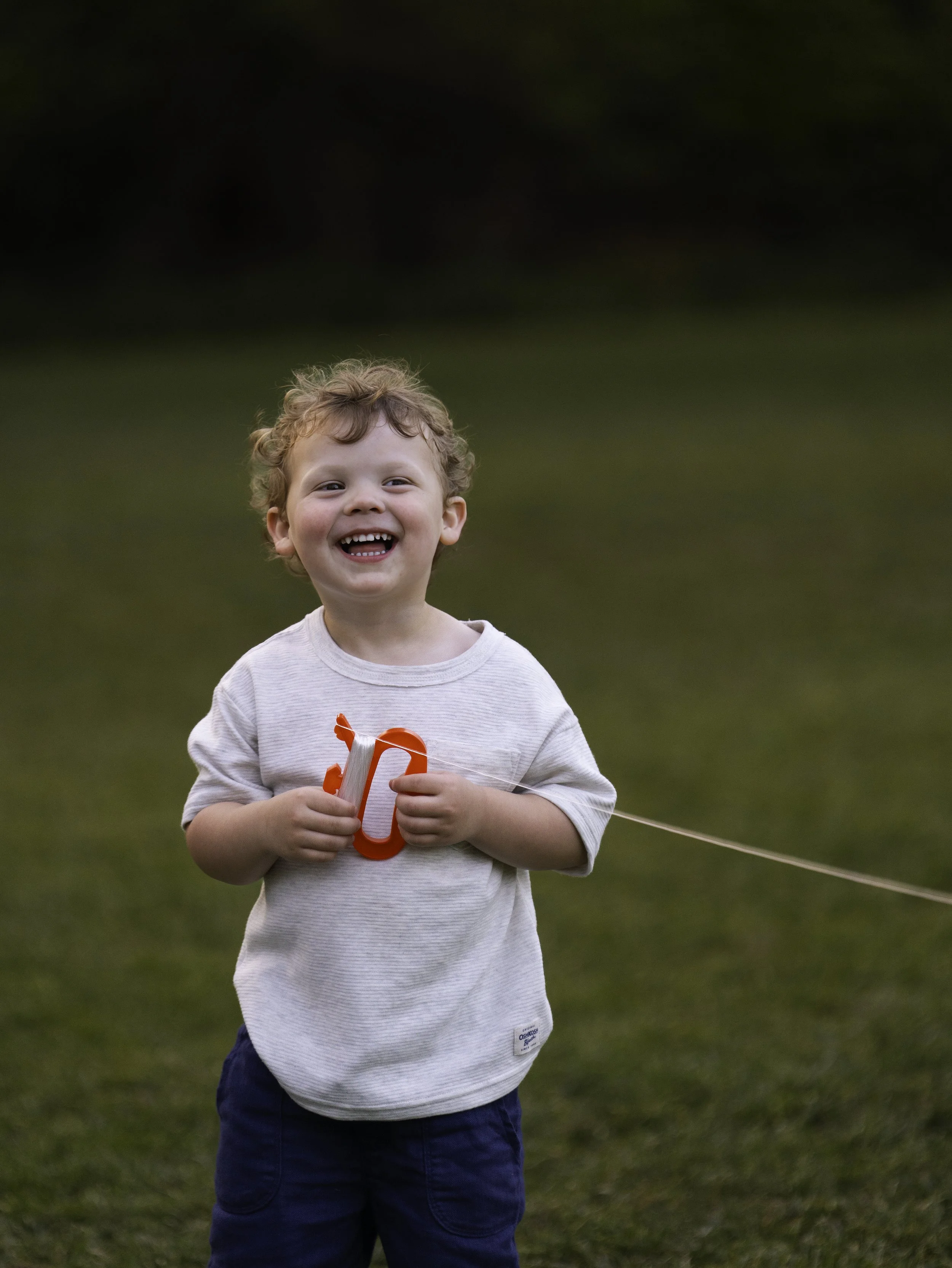 A young boy with curly blonde hair smiling and holding a red kite on a grassy field.