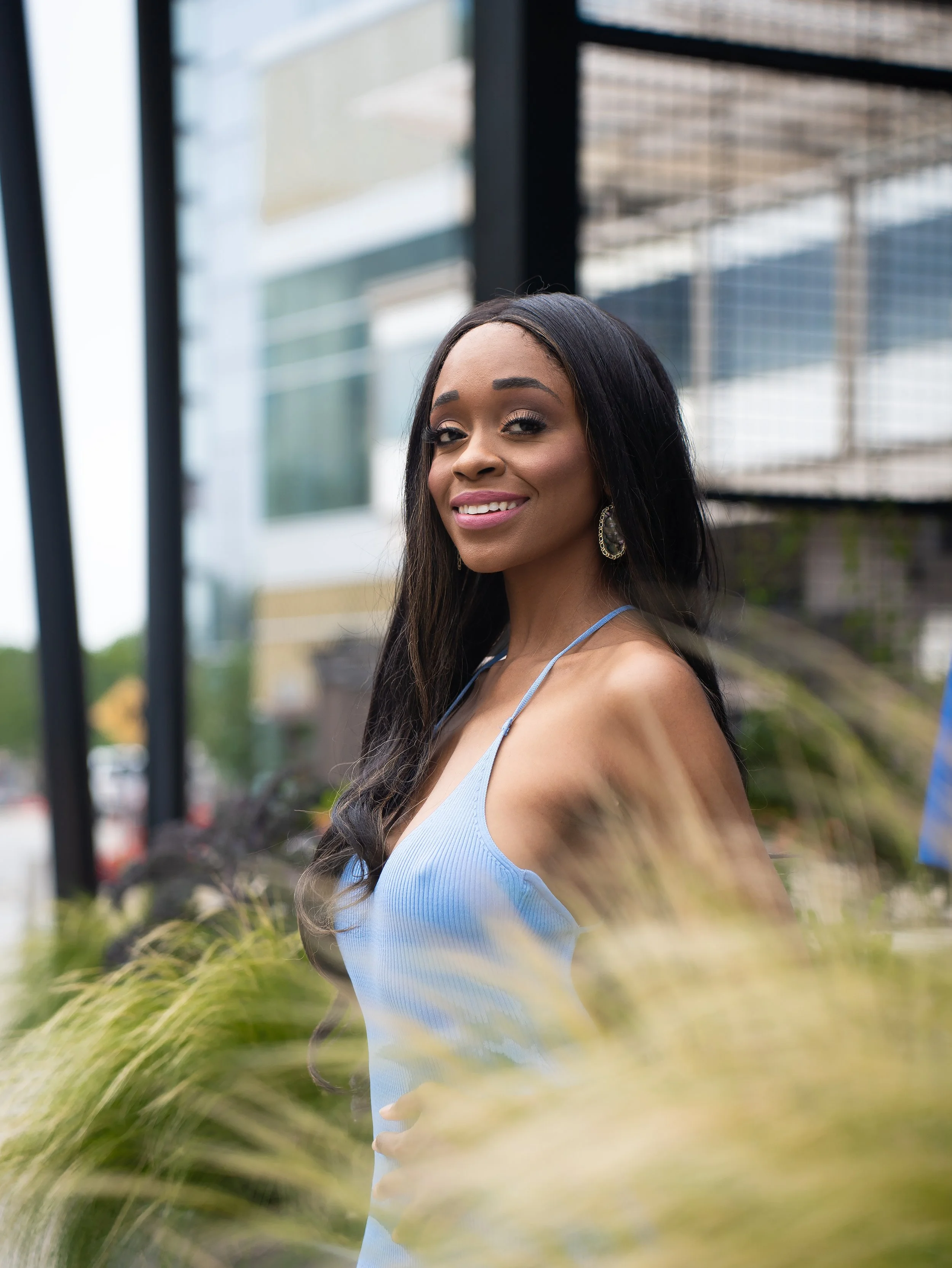 A woman with long black hair wearing a light blue spaghetti strap dress, smiling at the camera outdoors near a black metal fence and modern glass building.