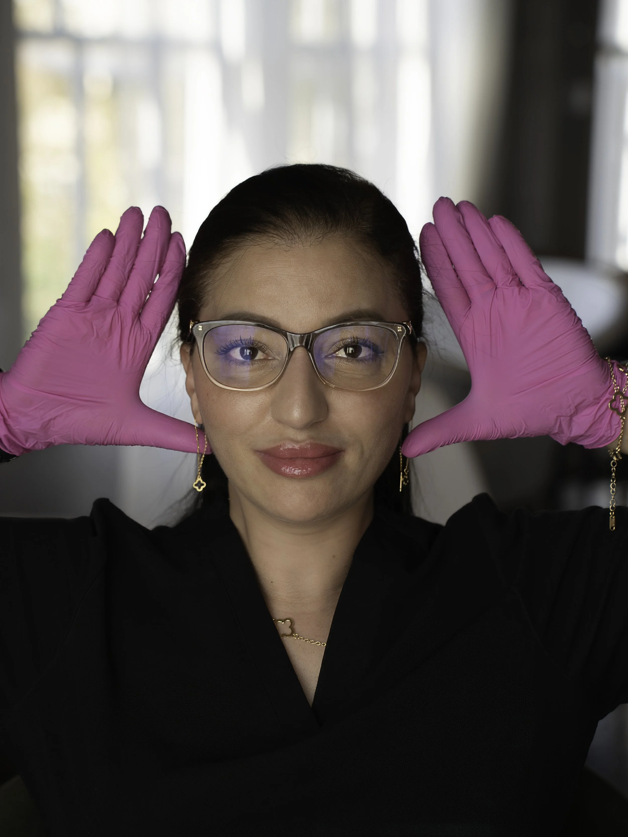 A woman wearing glasses, gold earrings, and a gold necklace, holding pink gloves to her head, with a neutral expression, in a room with curtains in the background.