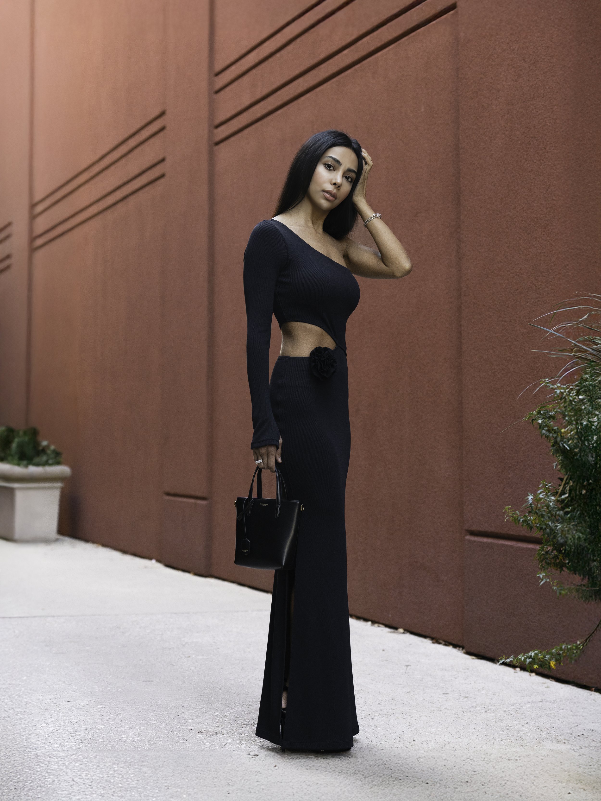 A woman in a long black evening gown standing outdoors against a red wall.