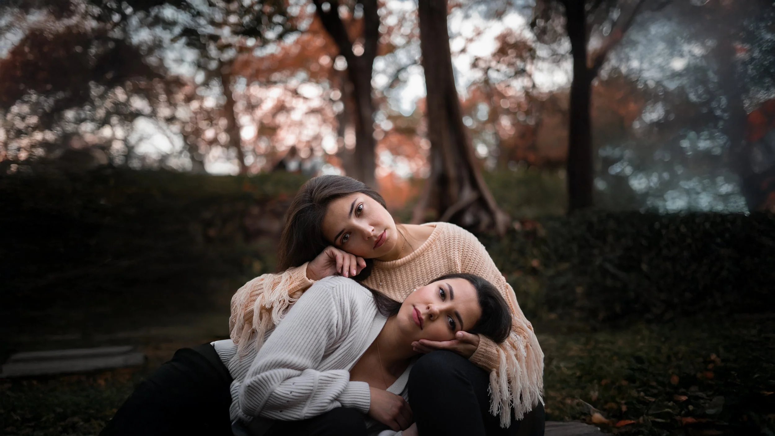 Two young women lying on the ground outdoors, with autumn trees in the background. One woman is resting her head on the other's shoulder, both looking at the camera.