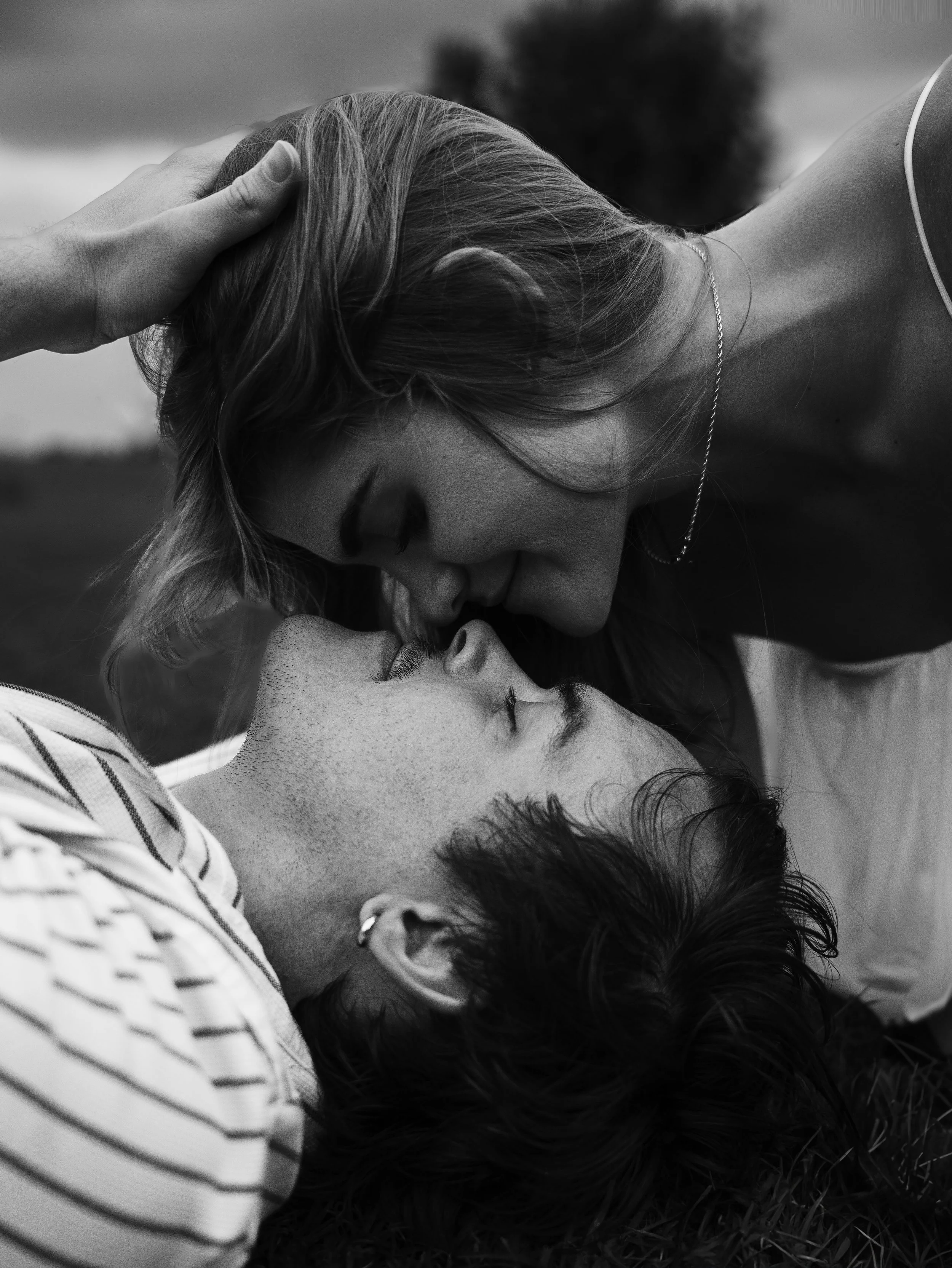 A black and white photo of a couple lying on the grass, close together. The woman is gently kissing the man's forehead while a hand softly touches her hair. The scene conveys tenderness and intimacy.