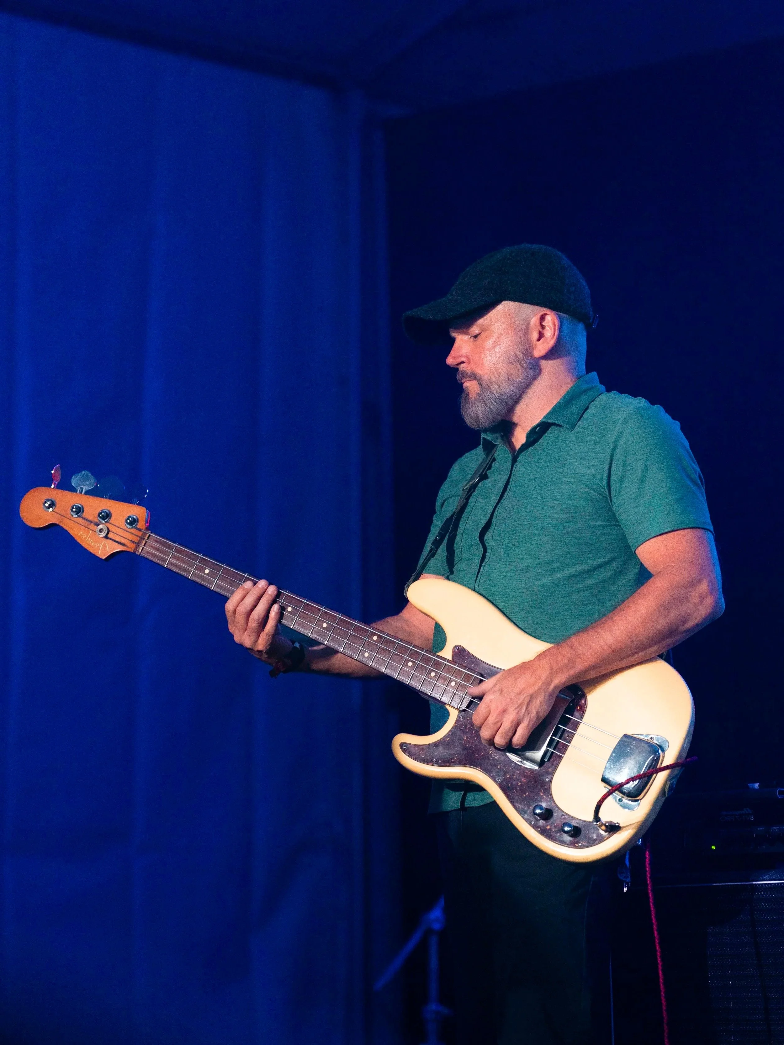 Male musician wearing a black cap and a green polo shirt playing an electric guitar on stage with a dark blue background.